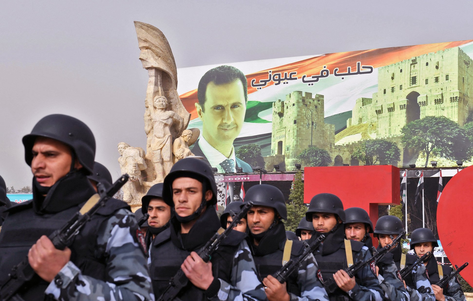 L'image montre un défilé militaire, avec des soldats en uniforme portant des casques et tenant des armes. En arrière-plan, on peut voir une grande affiche représentant un dirigeant, avec des bâtiments historiques et des éléments du drapeau syrien. La scène évoque une ambiance de démonstration de force et de soutien au gouvernement. Des sculptures monumentales sont également visibles, ajoutant une dimension artistique à l'ensemble.
