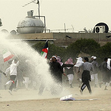 L'image semble montrer une scène de manifestation ou de rassemblement. On peut voir un groupe de personnes courant, certains tenant des drapeaux. Il y a également un jet d'eau qui pourrait provenir d'un canon à eau, suggérant une intervention des forces de l'ordre. L'arrière-plan comporte des bâtiments et un environnement rural, avec de la poussière soulevée par le mouvement. L'atmosphère semble tendue.