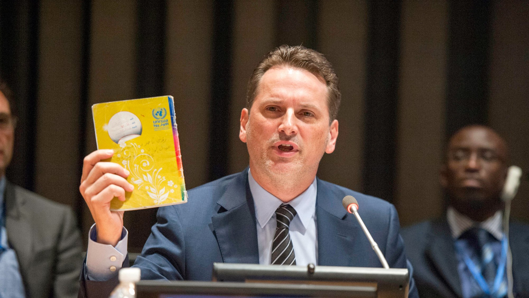 The image shows a man speaking at a podium during a conference or meeting, likely at a United Nations event. He is holding up a yellow booklet or document, which appears to be significant to his presentation. The nameplate in front of him reads "PIERRE KRAHENBUHL." In the background, there are several other individuals seated, and the setting has a formal atmosphere typical of international discussions. The man is engaged and emphasizing a point while addressing the audience.