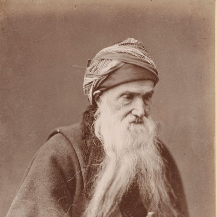 L'image montre un homme âgé avec une longue barbe blanche. Il porte un turban ou une coiffe enroulée autour de la tête, ainsi qu'une cape sombre. Son regard est concentré et il tient une canne. L'arrière-plan est neutre, mettant en valeur son visage et son expression, ce qui donne une impression de sagesse et de dignité.