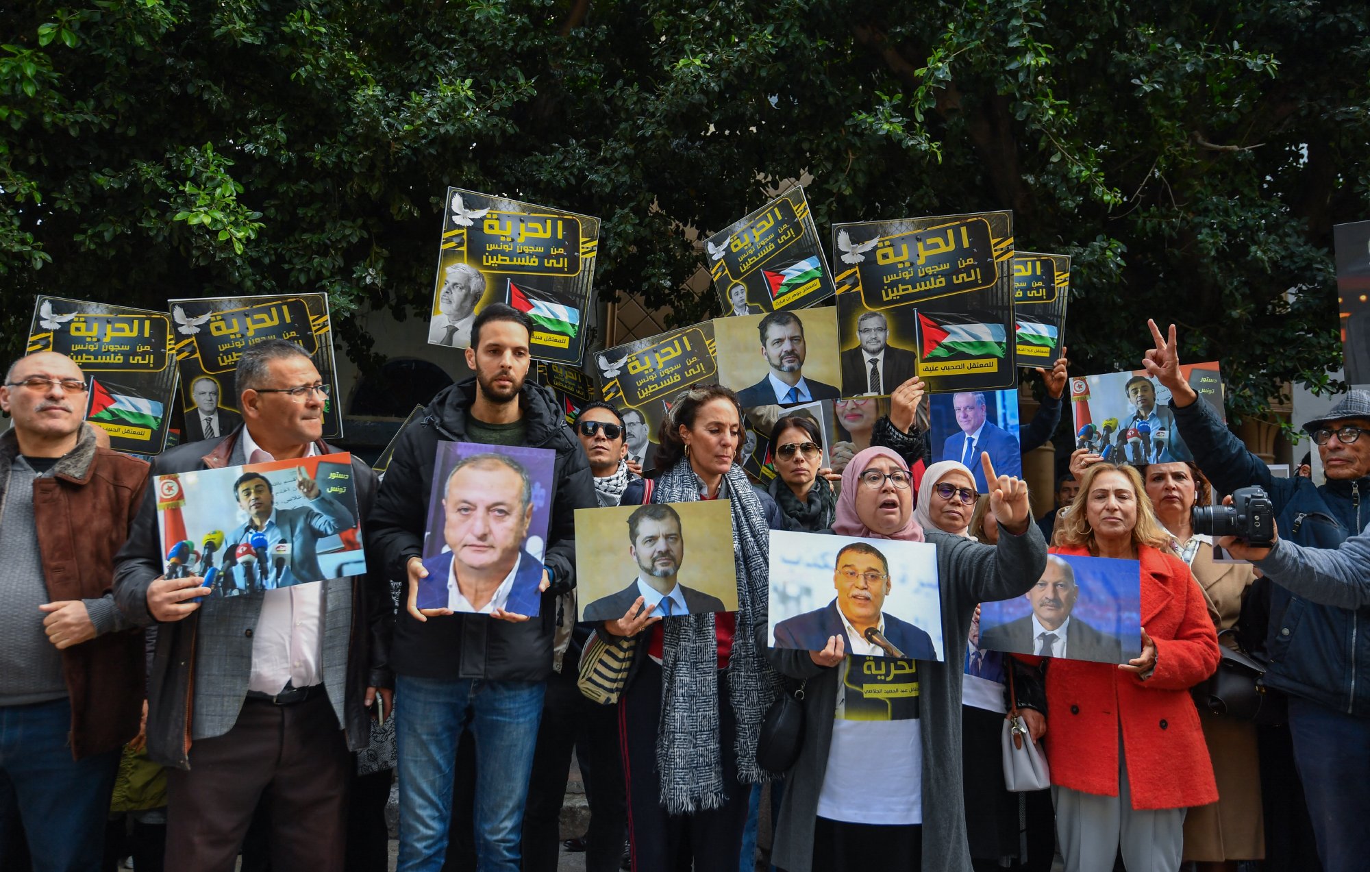 L'image montre un groupe de personnes réunies lors d'une manifestation. Elles tiennent des pancartes et des portraits de personnalités, exprimant des messages de soutien. L'ambiance est dynamique, avec des visages souriants et engagés.