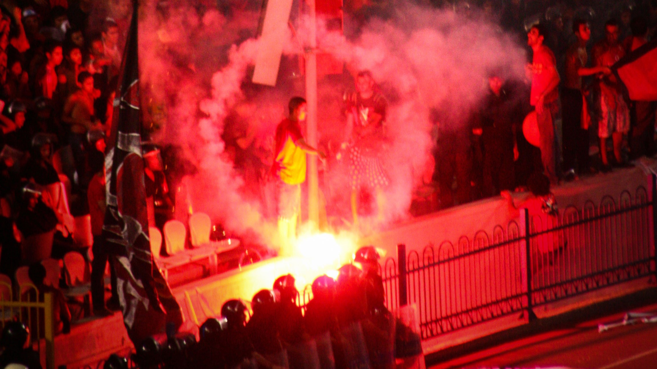 L'image montre une scène de manifestation avec une atmosphère intense. On peut voir des fumigènes rouges créant une nuée de fumée, ainsi que des personnes rassemblées autour d'une structure ou d'un symbole marquant, probablement dans un contexte de protestation. Au premier plan, une file de policiers en uniformes semble faire face aux manifestants. L'ensemble de la scène transmet une impression de tension et d'engagement émotionnel.