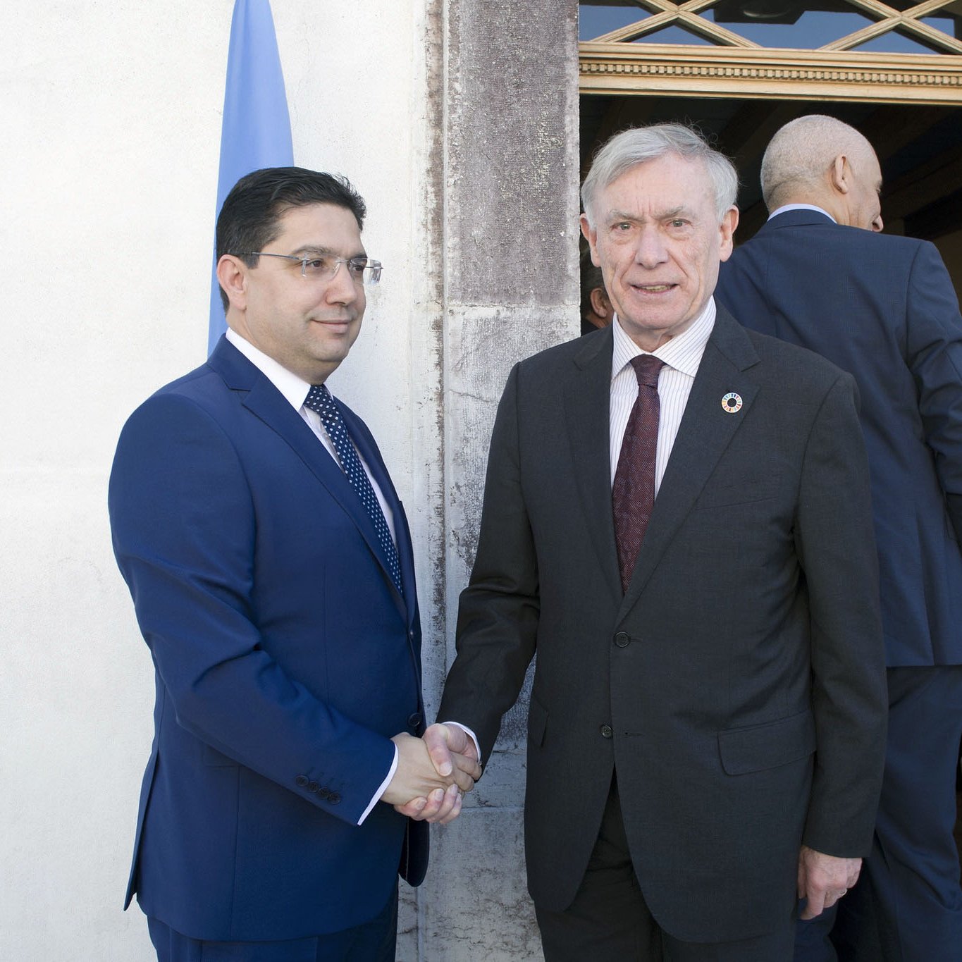 الصورة تظهر شخصين يتصافحان خارج مبنى. الشخص الأول يرتدي بدلة زرقاء ويبدو عليه الجدية. الشخص الثاني يرتدي بدلة داكنة ويظهر تعبيراً ودياً. في الخلفية، يمكن رؤية بعض الأشخاص الآخرين يدخلون أو يغادرون المكان، بالإضافة إلى العلم الذي يظهر في الزاوية.