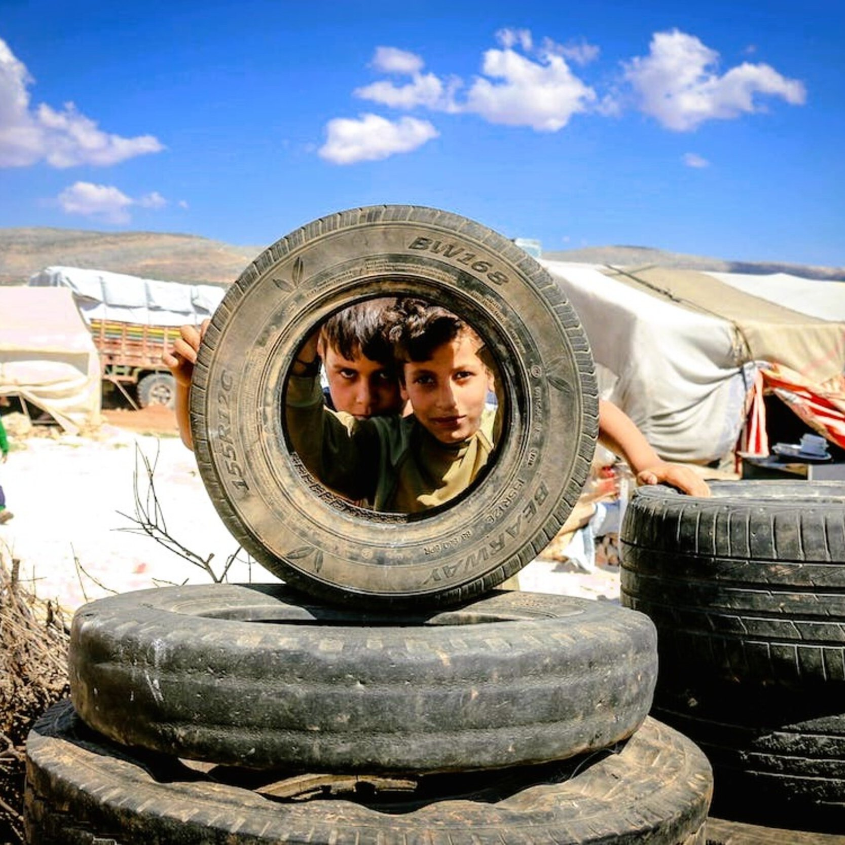 Nell'immagine si vedono due bambini che si affacciano attraverso un copertone di gomma, posto in orizzontale. I bambini, che sembrano curiosi e giocosi, sono circondati da altri copertoni impilati. Sullo sfondo si possono intravedere tende bianche e un paesaggio collinare sotto un cielo blu con alcune nuvole. L'atmosfera sembra essere di gioco e scoperta, nonostante l'ambiente circostante possa suggerire condizioni di vita difficili.