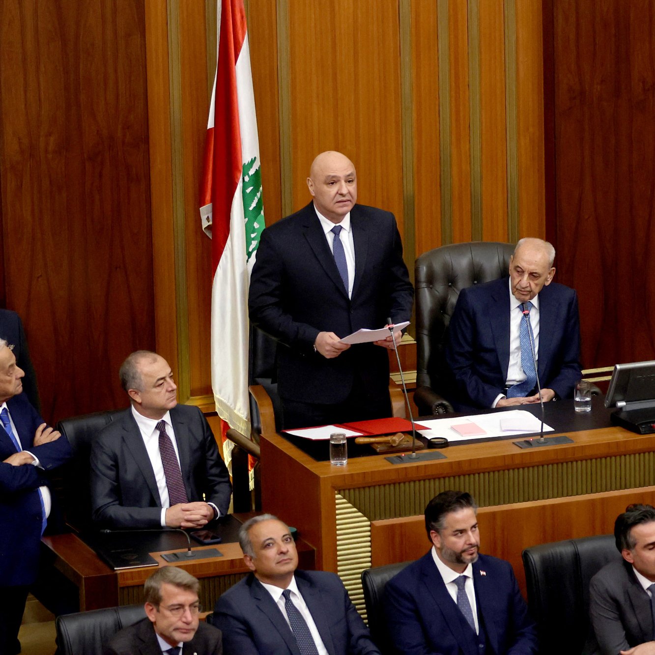 L'image présente une scène politique au sein d'un parlement. Au centre, un homme se tient debout, tenant des documents, probablement en train de s'adresser aux autres membres. À sa droite, un homme est assis derrière un bureau, semblant être un président ou un responsable de la séance. Plusieurs personnes sont debout et assises autour, portant des costumes, et affichent diverses expressions. En arrière-plan, on aperçoit le drapeau libanais. L'ambiance semble formelle et sérieuse, typique des assemblées législatives.