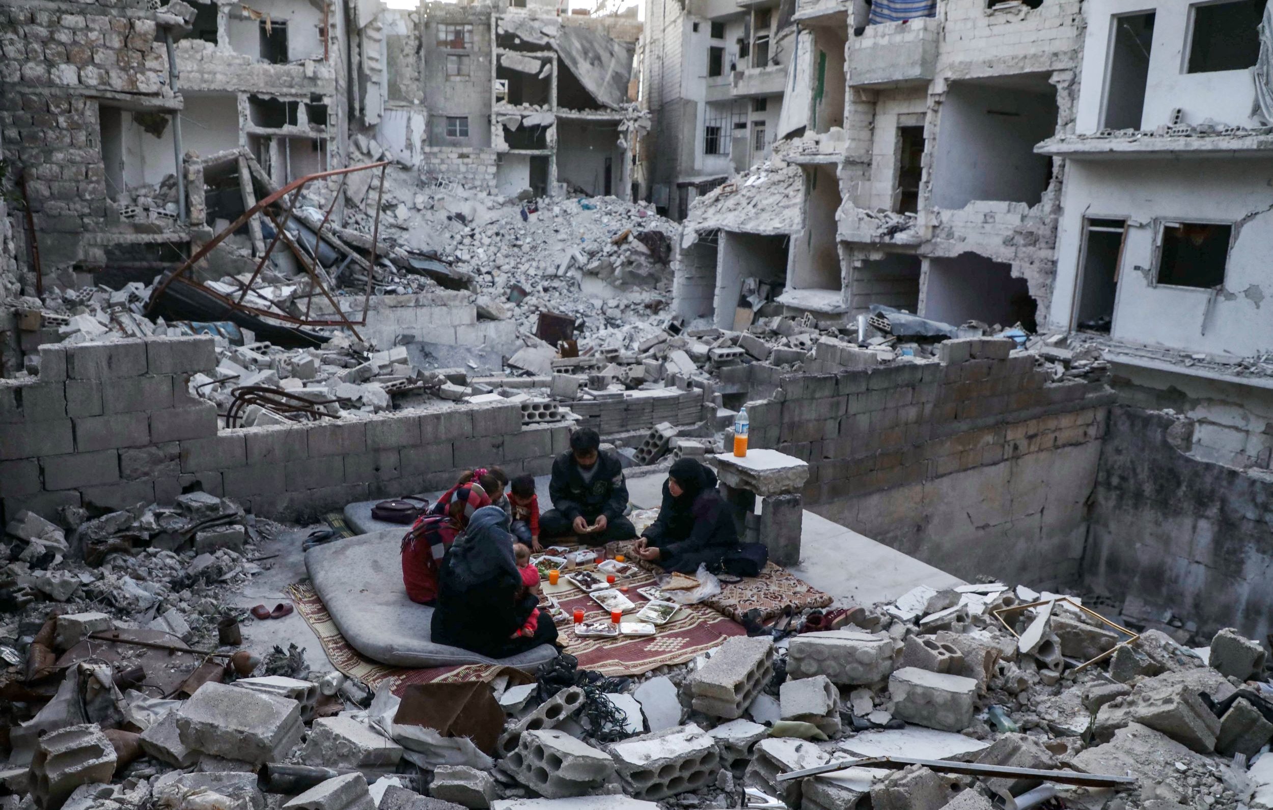 L'immagine mostra un contesto di distruzione, probabilmente in seguito a un conflitto. Al centro, un gruppo di persone è seduto su un tappeto, apparentemente intenti a giocare o a trascorrere del tempo insieme. Attorno a loro ci sono macerie e detriti di edifici crollati, con pareti distrutte e strutture parzialmente in piedi. La scena trasmette un senso di resilienza nel mezzo della devastazione.