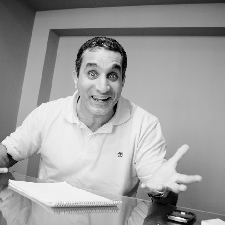 L'image montre un homme assis à une table, avec un air enthousiaste et des gestes expressifs. Il semble parler avec passion, ses bras levés et son visage affichant une grande animation. On peut voir des notes et un petit objet sur la table, suggérant qu'il est en train de discuter ou de partager ses idées. L'image est en noir et blanc, ce qui ajoute une dimension artistique à sa présentation.