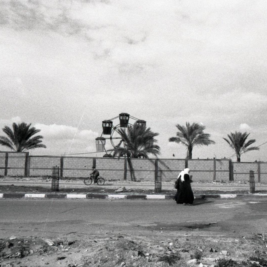 L'immagine ritrae una scena in bianco e nero che mostra una figura solitaria che cammina lungo una strada. Sullo sfondo, si possono vedere alcune palme e una ruota panoramica, che suggerisce la presenza di un parco o di un'area ricreativa. La composizione offre un'atmosfera malinconica e riflessiva, accentuata dalla scelta dei toni scuri e dalla mancanza di colori vivaci. La figura, vestita con abiti tradizionali, sembra essere in un momento di solitudine all'interno di un paesaggio urbano.