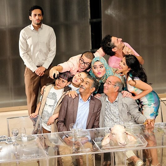 L'image montre un groupe de personnes rassemblées autour d'une table, apparemment lors d'un événement convivial. Il y a des adultes et des enfants, certains prenant la pose pour un selfie. Les expressions sont joyeuses et décontractées, avec des sourires sur presque tous les visages. À l'arrière-plan, un homme se tient debout, regardant la scène. La table est décorée avec des verres et un plat, ajoutant une ambiance festive à la scène. L'ensemble dégage une atmosphère d'amitié et de bonheur partagé.