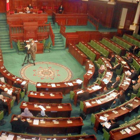 L'image montre l'intérieur d'un parlement, probablement un hémicycle de l'assemblée nationale. On peut voir plusieurs rangées de sièges verts disposés en arc de cercle, où des membres semblent assister à une session. Au centre, un tapis décoratif est visible, et à l'arrière-plan, on aperçoit une estrade pour les orateurs. Des caméras sont présentes, indiquant que la session est probablement diffusée. L'ensemble dégage une atmosphère formelle et politique.
