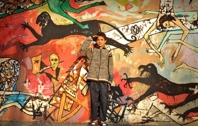 L'image montre un jeune garçon en train de faire un salut militaire devant un mur coloré, sur lequel sont peintes diverses figures stylisées et abstraites. Le mur présente des scènes dynamiques, avec des motifs de monstres et d'êtres humains, créant un contraste frappant avec le garçon qui porte un manteau gris et un pantalon sombre. Son expression et sa posture semblent témoigner de confiance ou de détermination. Les couleurs vives et les formes exagérées de la fresque attirent l'œil et ajoutent une dimension artistique à la scène.