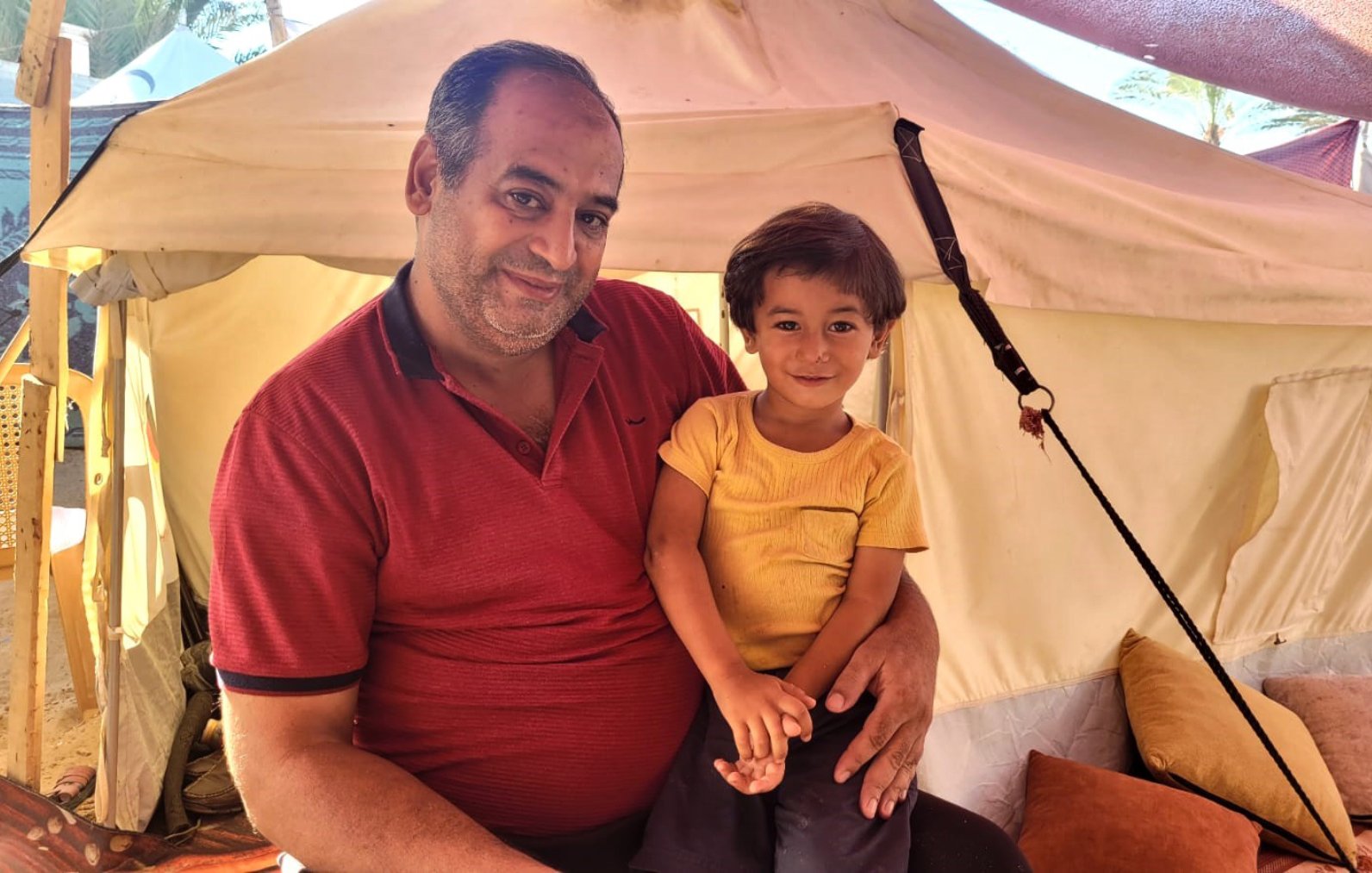 L'image montre un homme souriant assis à l'intérieur d'une tente, tenant un jeune enfant sur ses genoux. L'homme porte une chemise rouge et a l'air détendu et joyeux. L'enfant, qui porte un t-shirt jaune, sourit également et semble heureux. L'intérieur de la tente est simple, avec des coussins au sol, créant une atmosphère conviviale et chaleureuse.