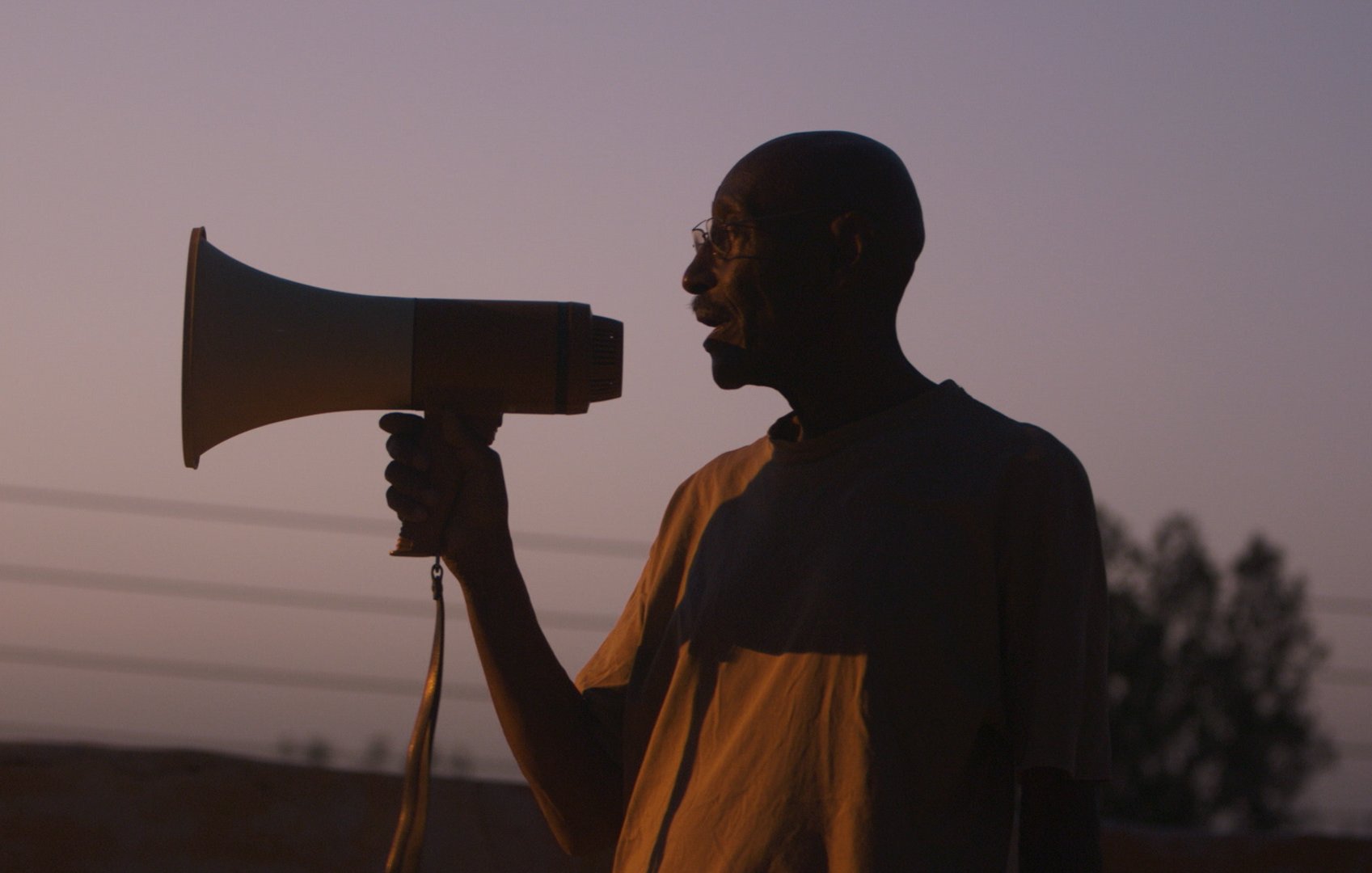 رجل يحمل مكبر صوت، silhouetted أمام غروب الشمس، يبدو وكأنه ينادي بشيء مهم.