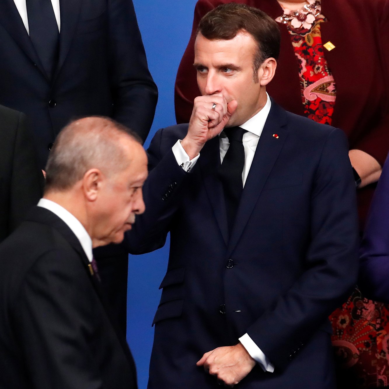 L'image montre un groupe de dirigeants politiques rassemblés lors d'un événement officiel. Au premier plan, on peut voir un homme qui se tient à gauche, probablement en train de parler ou d'écouter. Au centre, un homme en costume sombre semble réfléchir, avec une main devant sa bouche. À droite, une femme porte un vêtement violet et observe la scène avec un léger sourire. L'atmosphère semble être formelle, typique des rencontres diplomatiques.