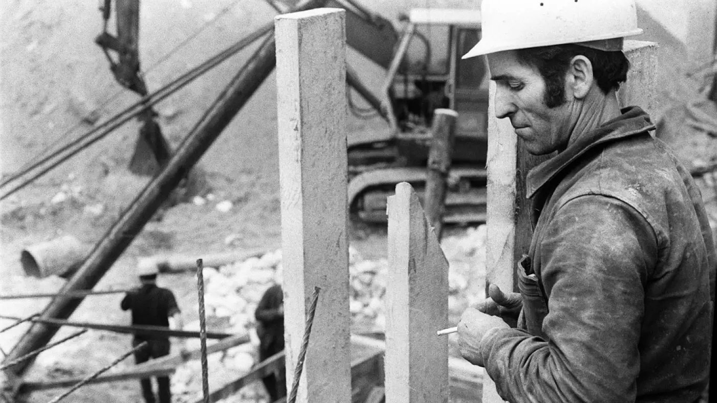 في الصورة، يظهر عامل بناء يرتدي خوذة بيضاء وهو يقف بالقرب من مجموعة من الأعمدة الخرسانية. يبدو أنه مركز على عمله، حيث يمسك بيده سيجارة. في الخلفية، يوجد عمال آخرون وأحدهم يعمل على آلة ثقيلة تُستخدم في البناء. الجو العام للصورة يتسم بالجدية والتركيز، ويظهر البيئة البنائية المحيطة والتي تشمل الأتربة والمواد الإنشائية.