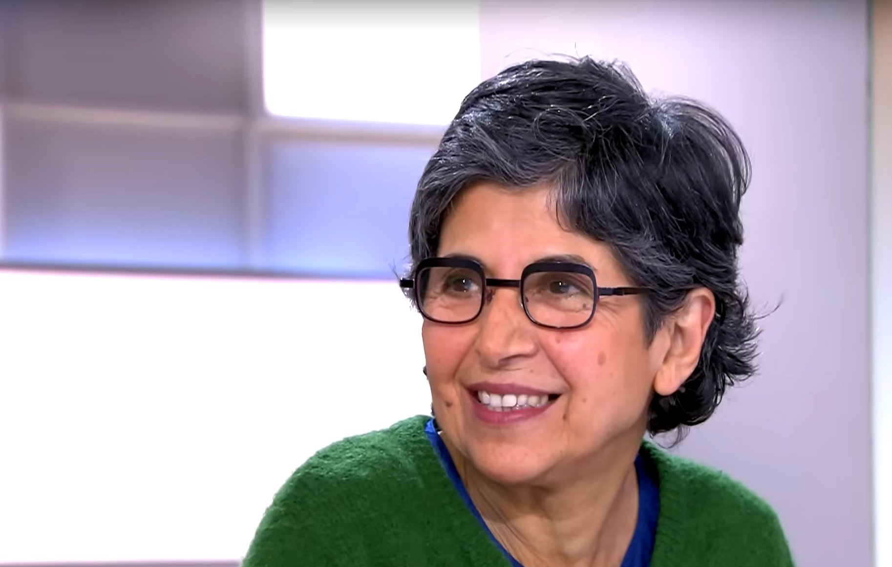 L'image montre une femme souriante avec des cheveux gris et courts. Elle porte des lunettes et un pull de couleur verte. Son expression est amicale et chaleureuse, indiquant qu'elle participe à une conversation. L'arrière-plan est flou, suggérant qu'elle se trouve dans un environnement de studio ou d'interview.