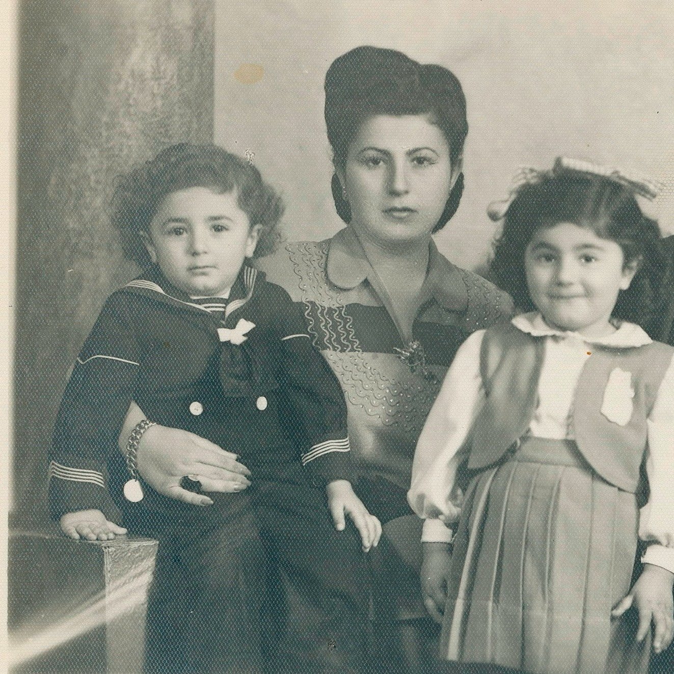 Cette image en noir et blanc montre une famille posant ensemble. À gauche, un jeune garçon portant une tenue de marin est assis. À côté de lui, une femme au style classique avec des cheveux coiffés en pompadour se tient. En face d'elle, une petite fille avec un sourire doux porte une robe avec un gilet. À droite, un homme en costume, portant des lunettes, regarde la caméra. L'arrière-plan est sobre, ajoutant une touche nostalgique à cette photo de famille.