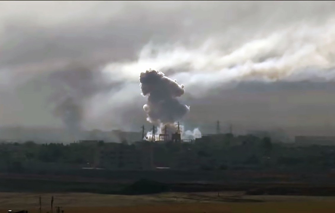 La imagen muestra una escena con humo denso y nubes oscuras que se elevan desde un área urbana. Se aprecian instalaciones industriales o edificios en el fondo, y el humo parece provenir de una explosión o un incendio. El cielo está nublado, lo que contribuye a una atmósfera sombría y tensa en la imagen.