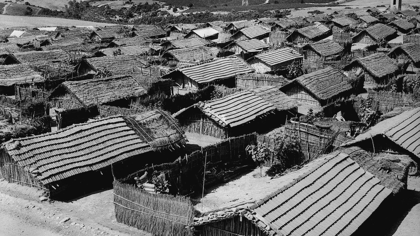 L'image montre un paysage rural avec plusieurs maisons en bois et en matériau naturel, probablement des huttes, disposées de manière dense. Les toits sont recouverts de paille ou d'un matériau similaire. On aperçoit également des collines verdoyantes à l'arrière-plan, ce qui suggère un environnement rural. Le ciel est clair, et l'ensemble de la scène a une tonalité noire et blanche, conférant une atmosphère nostalgique et ancienne à l'image.