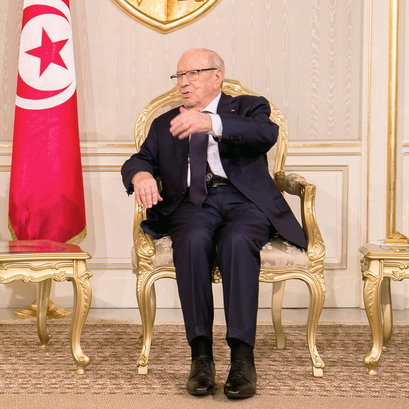 L'image montre un homme assis sur une chaise en or dans un environnement intérieur élégant. Il porte un costume sombre et des lunettes. À sa gauche, il y a un vase avec des fleurs blanches. Derrière lui, on aperçoit le drapeau tunisien. L'homme semble être en train de parler ou de gesticuler, indiquant une conversation ou une discussion en cours. Les murs et le décor environnant évoquent une atmosphère formelle et institutionnelle.