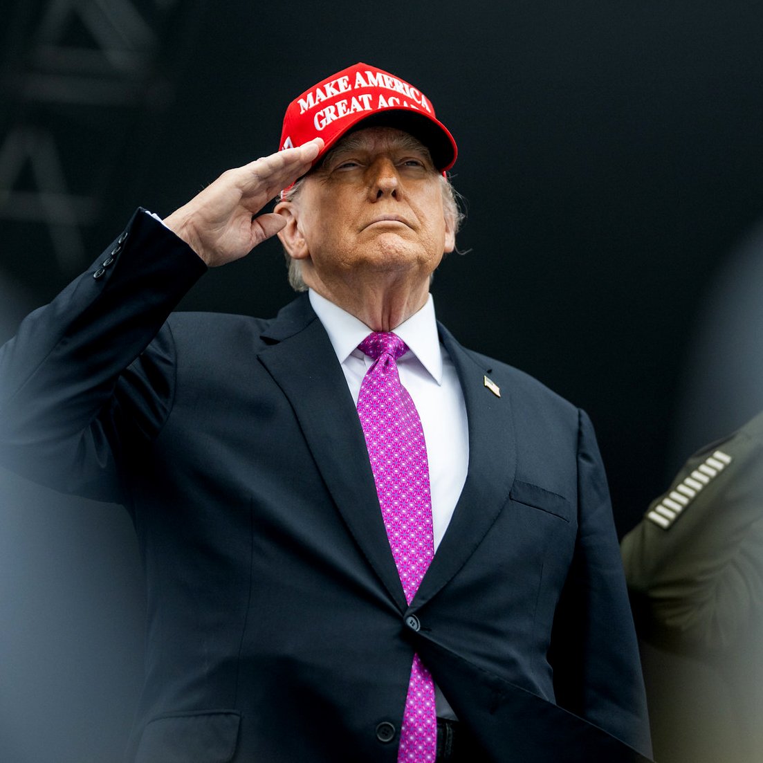 Homme en costume noir, portant une casquette rouge, effectuant un salut.