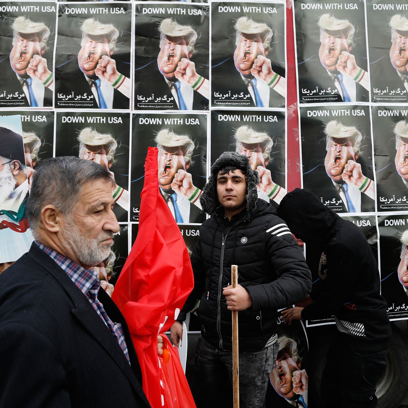 L'image montre une manifestation avec des affiches en arrière-plan affichant des personnages aux visages provocateurs, probablement des figures politiques, portant des coiffures spécifiques. Des gens tiennent des pancartes et des drapeaux, tandis qu'un homme sur le devant semble exprimer un message clair de désaccord ou de protestation. L'ensemble de la scène dégage une ambiance de contestation, soulignant des tensions politiques.