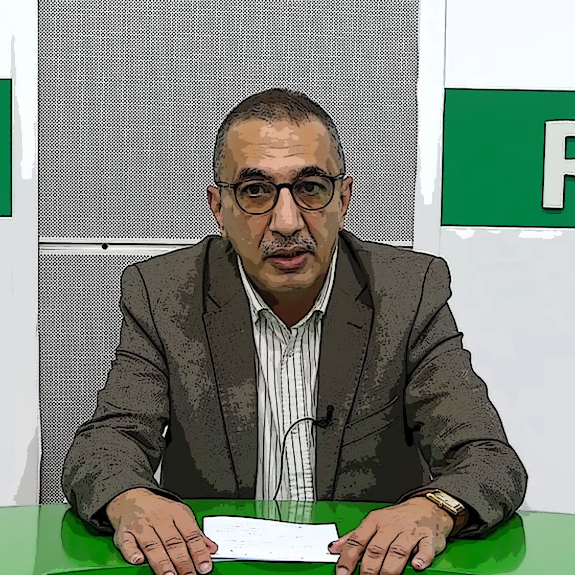 L'image montre un homme devant un bureau, probablement dans un studio de télévision ou de radio. Il porte des lunettes et un costume, avec une chemise rayée. Le bureau est vert, et il semble tenir des documents ou un script. En arrière-plan, il y a des éléments graphiques avec les mots "RADIO" et une autre inscription non visible. L'homme a une expression sérieuse et semble prêt à faire une présentation ou une annonce.