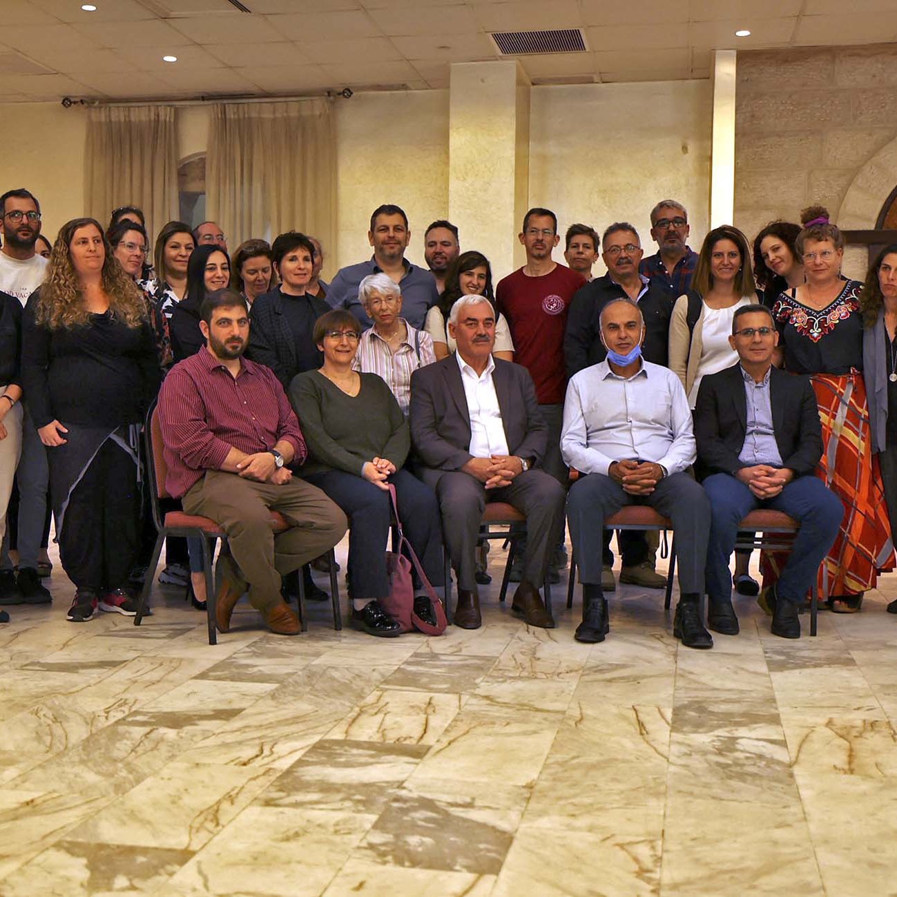 L'image montre un grand groupe de personnes rassemblées dans une salle, probablement pour un événement ou une célébration. Les gens sont assis et debout, souriant et regardant vers l'objectif. L'espace présente des murs en pierre et un sol en marbre, créant une atmosphère conviviale. On peut remarquer une variété de styles vestimentaires parmi les participants, ce qui indique une diversité au sein du groupe.