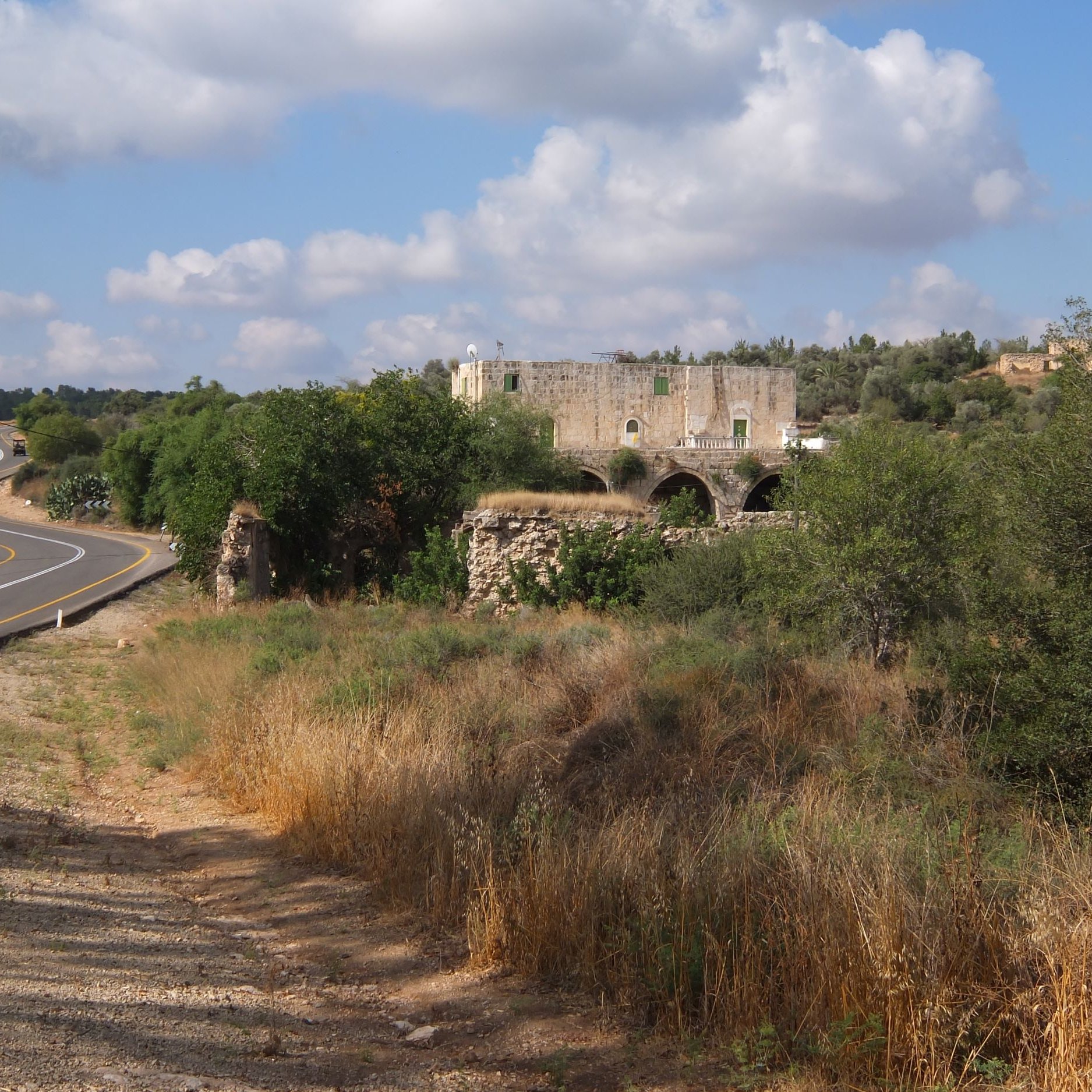 L'image montre un paysage rural avec une route sinueuse sur la gauche. À droite, il y a des herbes hautes et quelques arbres. Au fond, on aperçoit des ruines d'un bâtiment en pierre, probablement une ancienne construction, entourées de verdure. Le ciel est légèrement nuageux, créant une atmosphère paisible et naturelle.