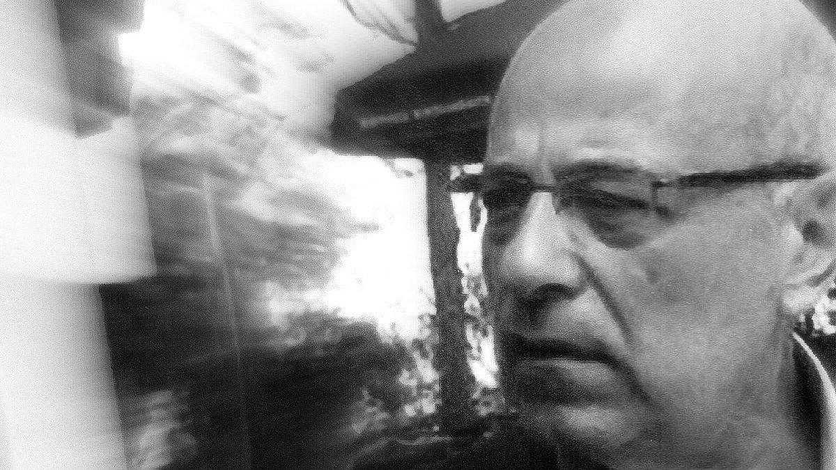 تظهر الصورة رجلًا في منتصف العمر، يبدو جادًا ومتأملًا. يعبر وجهه عن التفكير العميق أو الانشغال بشيء ما. يبدو أن الصورة قد أُخذت في مكان خارجي، حيث يمكن رؤية بعض العناصر الطبيعية أو المعمارية في الخلفية، مما يعطي إحساسًا بالعمق. الصورة تبدو مشوشة قليلاً، مما يضيف تأثيرًا فنيًا إليها.