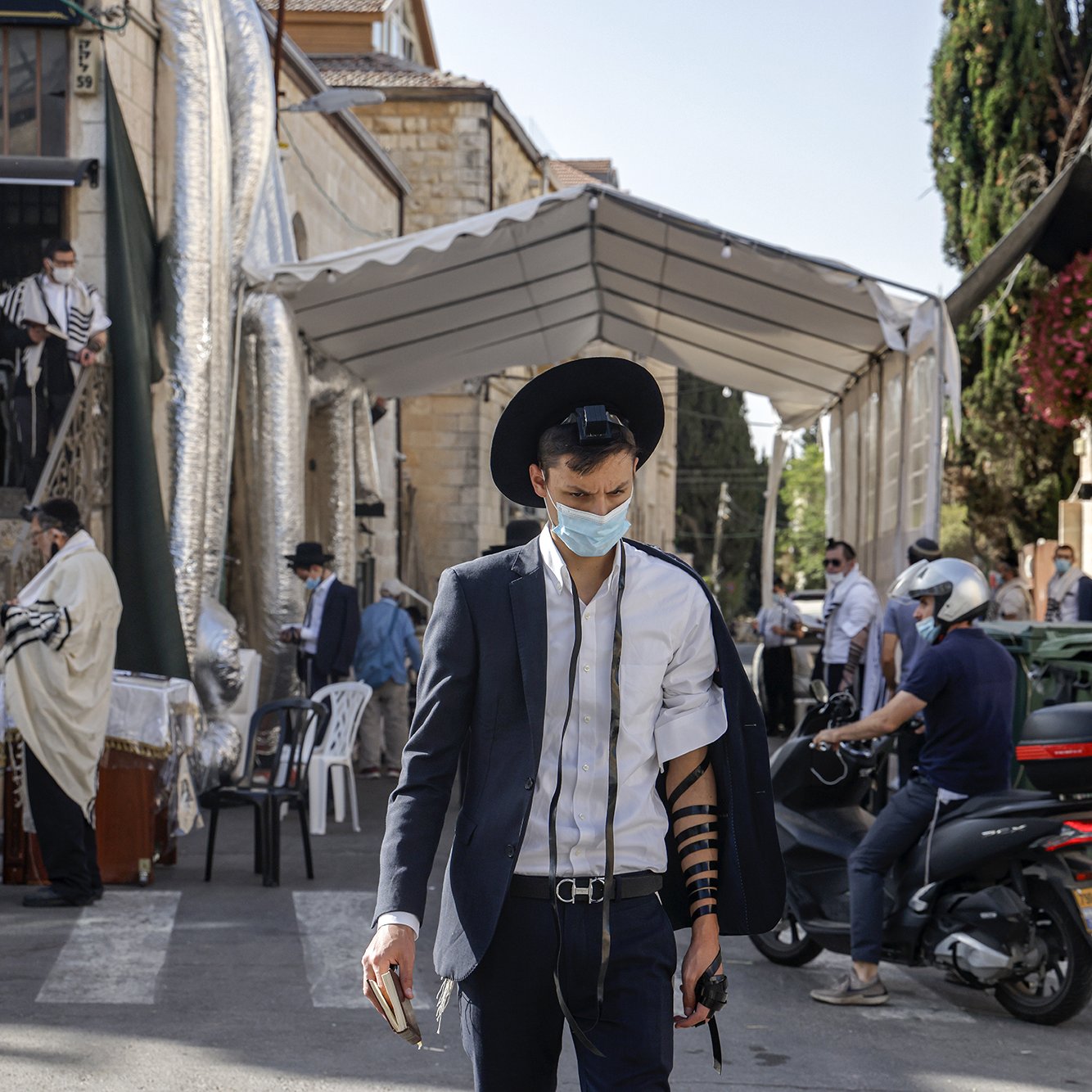 L'image montre un homme marchant dans une rue, portant un costume noir et un chapeau. Il a un masque facial et est vêtu d'un tefillin sur son bras. En arrière-plan, on peut voir un endroit semblant être un lieu de prière, avec des gens habillés en vêtements traditionnels. Des panneaux et de la végétation décorent le paysage urbain. La scène évoque une atmosphère communautaire et religieuse.