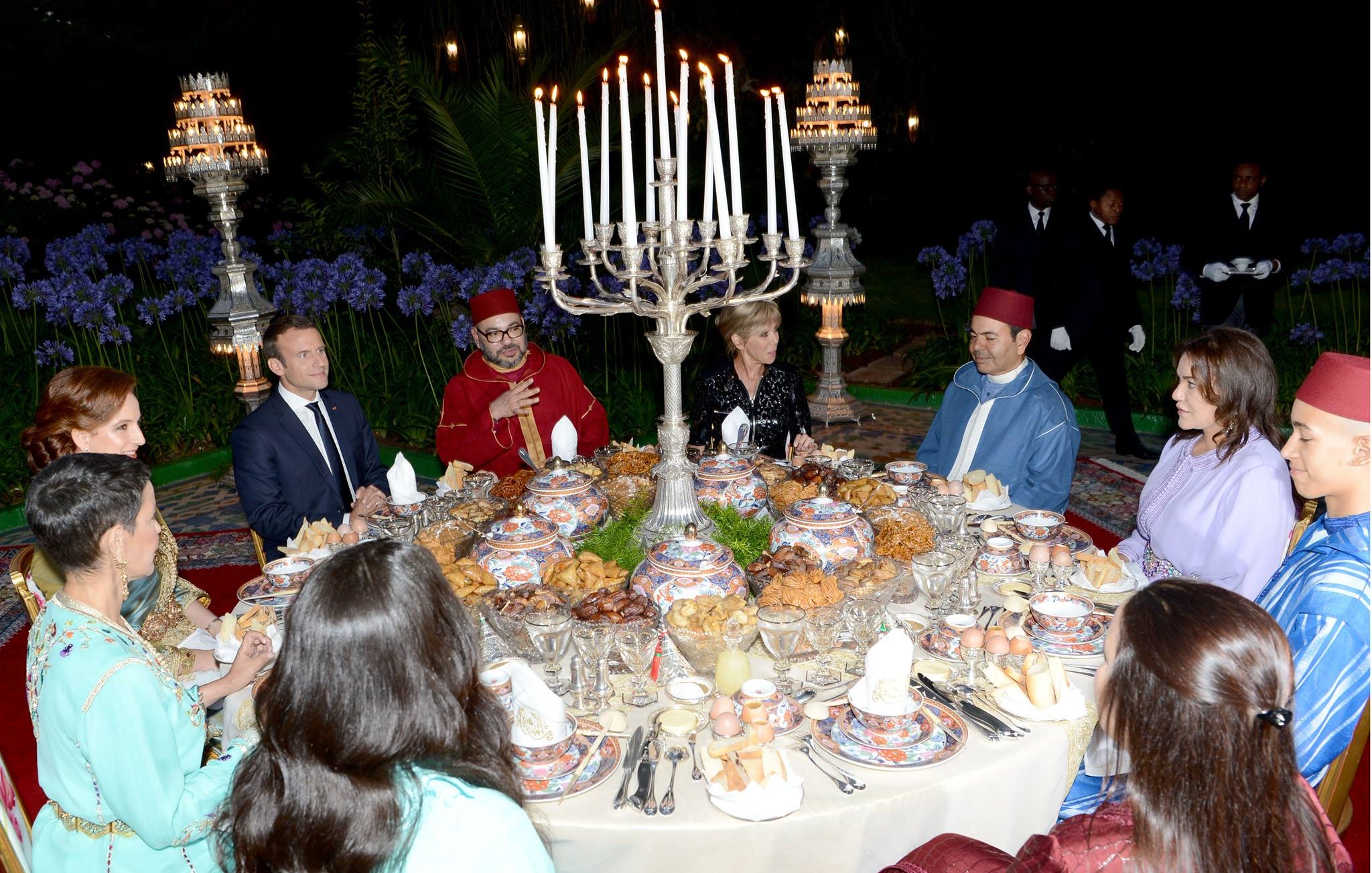 L'image montre une scène de dîner élégant, probablement dans un cadre extérieur. Une grande table est dressée avec une variété de plats et de délicieuses spécialités culinaires. De nombreuses bougies sont allumées, créant une ambiance chaleureuse et festive. Des convives, habillés en vêtements traditionnels, sont assis autour de la table, engageant des discussions animées. Au fond, on peut apercevoir des éléments décoratifs et des personnes en costume qui ajoutent à l'atmosphère formelle de l'événement.