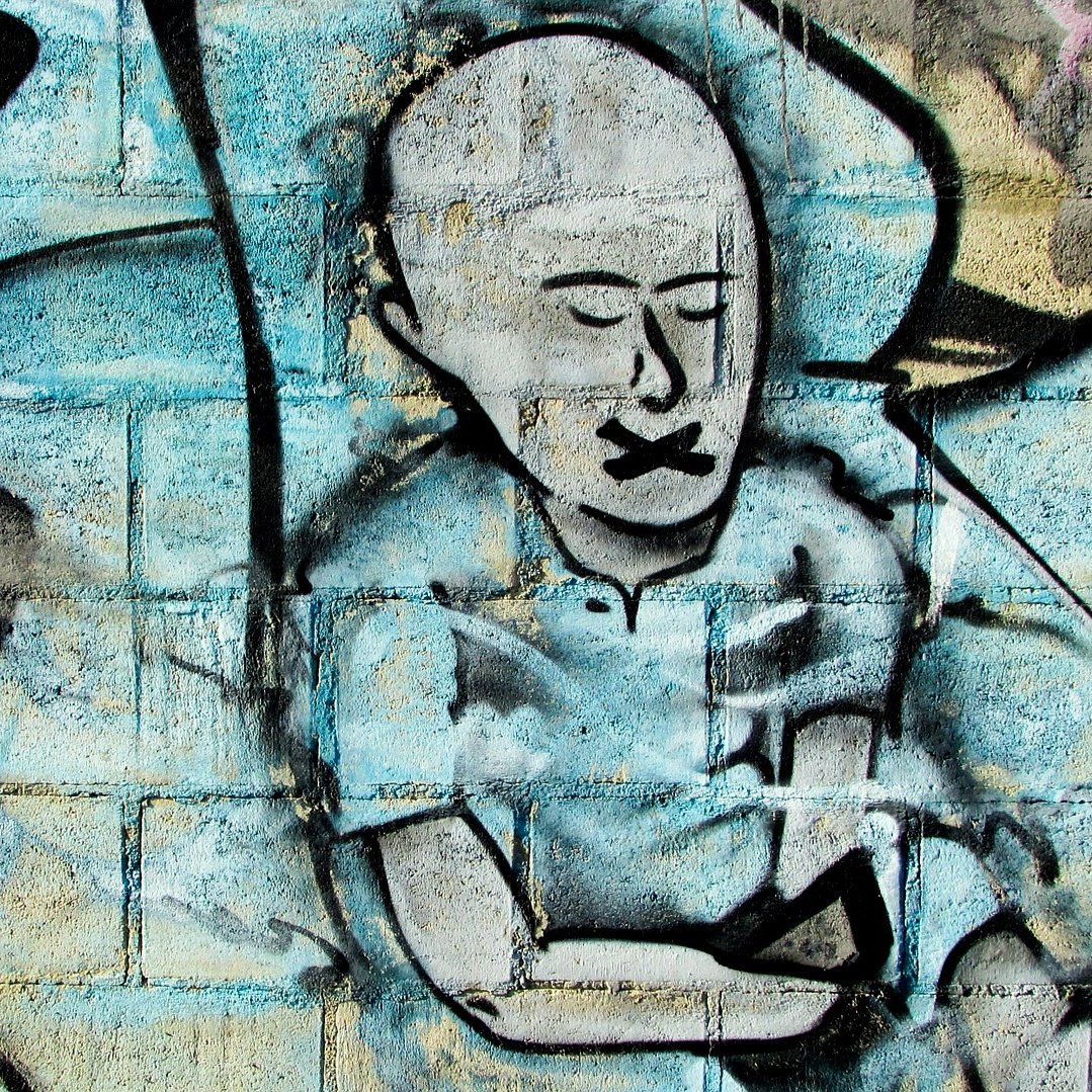 L'image présente un graffitti sur un mur. On y voit le dessin d'une figure humaine stylisée, sans cheveux, avec un visage exprimant le silence, probablement pour symboliser l'oppression ou le manque de voix. Ses lèvres sont représentées comme étant scellées. Le fond est composé de couleurs bleu et gris, avec des formes abstraites qui ajoutent de la texture à l'œuvre. L'ensemble dégage une ambiance à la fois poignante et réfléchie.