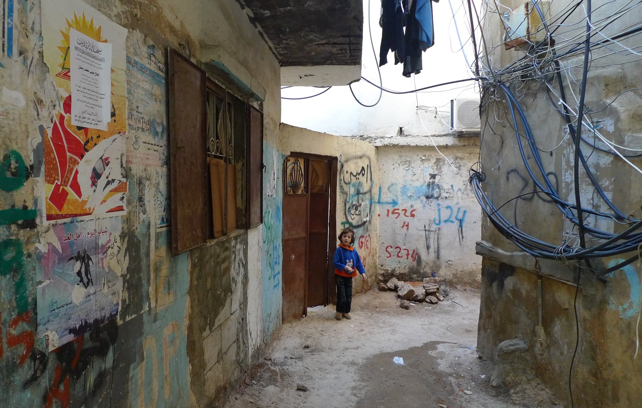 L'image montre une ruelle étroite et dégradée, bordée de murs en béton sur lesquels sont peints des graffitis et des affiches colorées. À gauche, une porte en bois est partiellement ouverte, tandis qu'à droite, on peut voir des fils électriques accrochés. Au fond de la ruelle, un jeune enfant se tient près de l'entrée, regardant vers l'objectif. L'atmosphère semble à la fois désolée et animée par les couleurs des murs et des affiches.