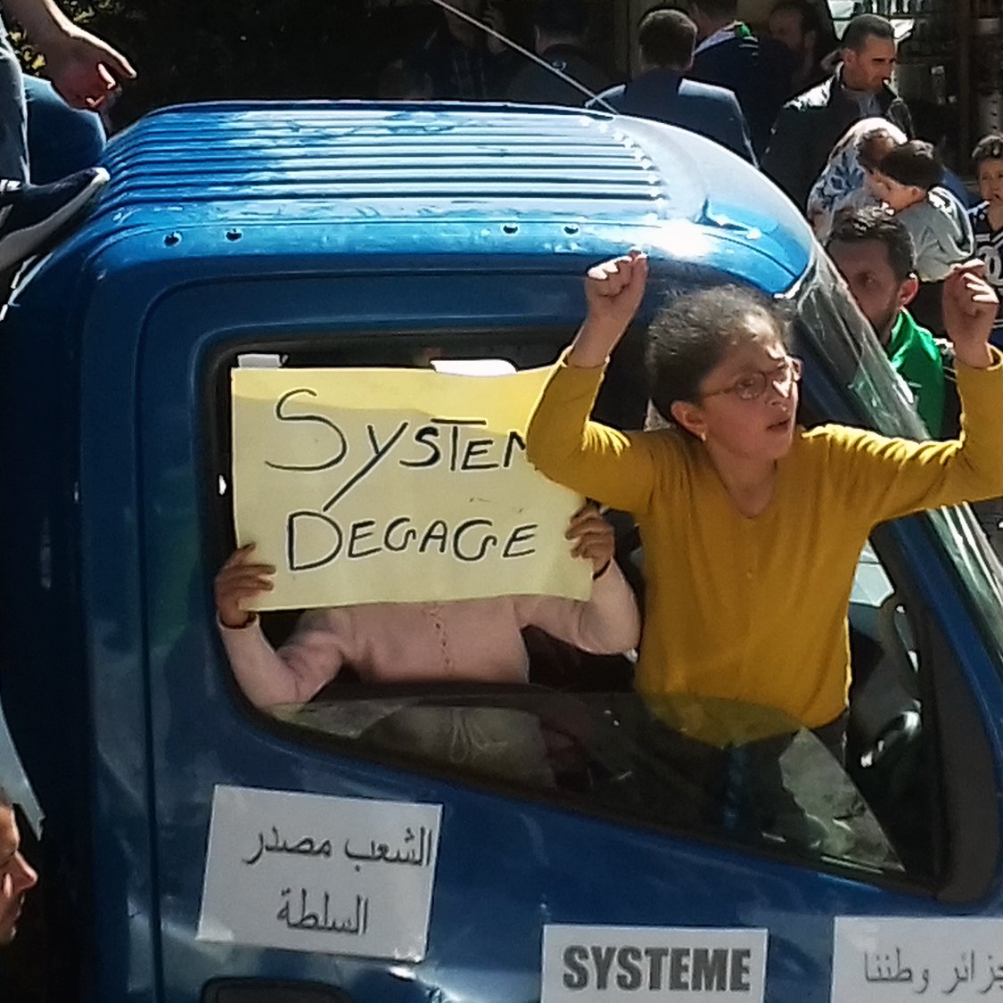 The image shows a group of people in a demonstration or protest, focused around a blue truck. One person is prominently displaying a sign that reads "Systeme Degage," indicating a call for the system to go or be removed. In the truck, another individual can be seen holding a sign. The atmosphere appears energetic, with participants expressing their views passionately. The surrounding crowd suggests a larger movement, likely addressing political or social issues.