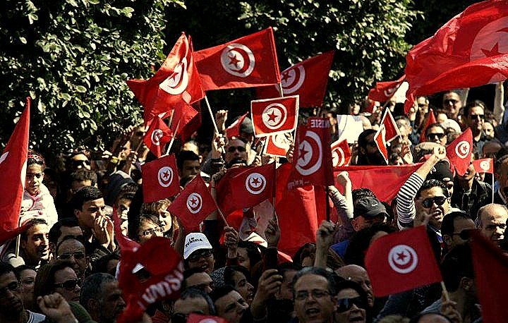 L'image montre une foule de personnes rassemblées, agitant des drapeaux tunisiens rouges avec un cercle blanc et une étoile à cinq branches. Les manifestants semblent animés, levant les bras en signe de solidarité ou de protestation. On peut voir un paysage urbain en arrière-plan avec des arbres, ce qui suggère un rassemblement en plein air. L'atmosphère semble énergique et engagée, reflétant un moment de mobilisation collective.