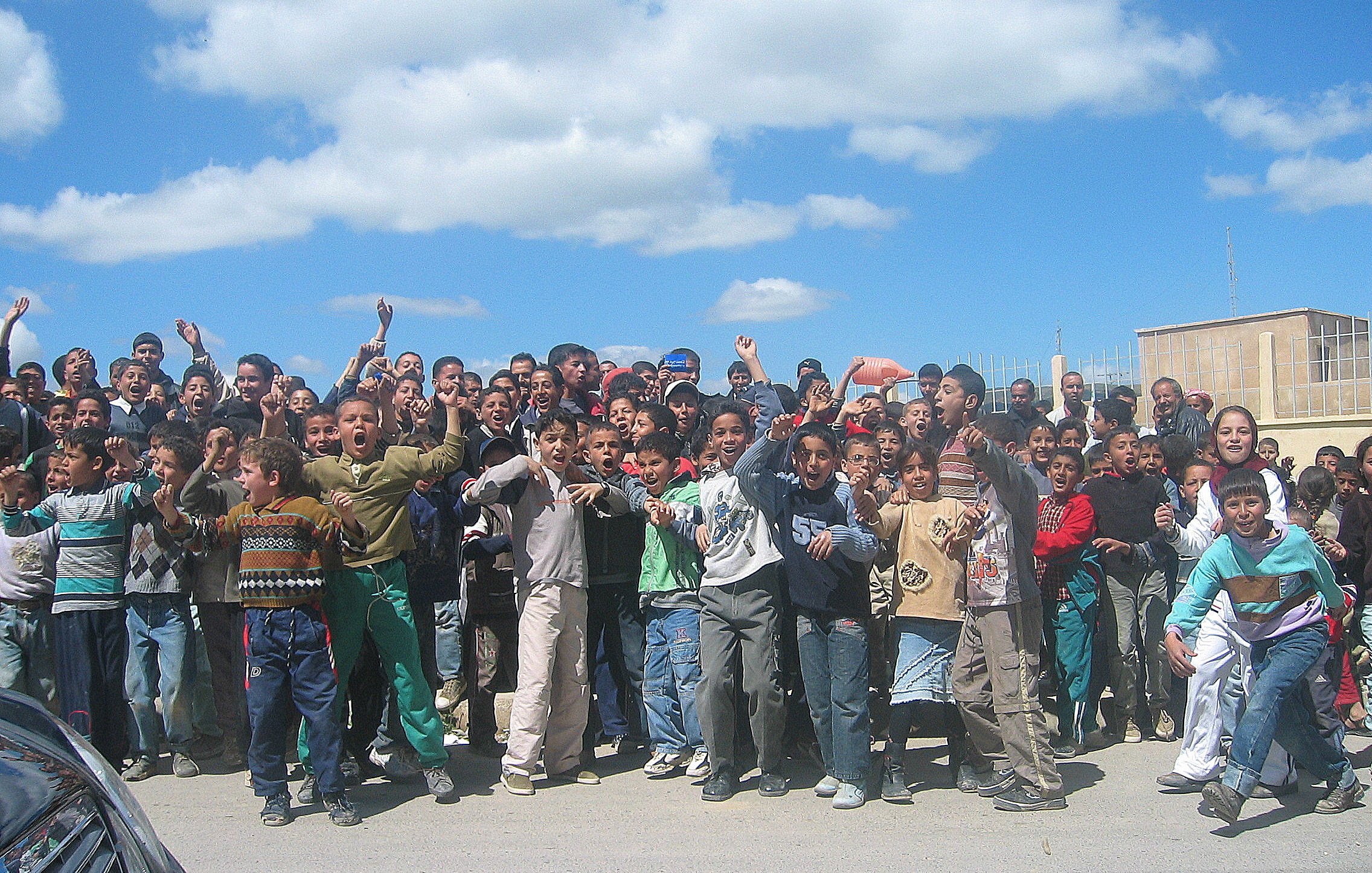 تظهر في الصورة مجموعة كبيرة من الأطفال والشباب يتجمعون في مكان واحد، يبدو أنهم متحمسون ويقومون بالتفاعل مع شيء ما، ربما يجتمعون لمتابعة حدث أو نشاط معين. السماء في الخلفية زرقاء مع بعض السحب، والمكان يبدو كأنه في منطقة حضرية. يرتدي الأطفال ملابس متنوعة، وتعكس تعبيراتهم الحماس والإثارة.