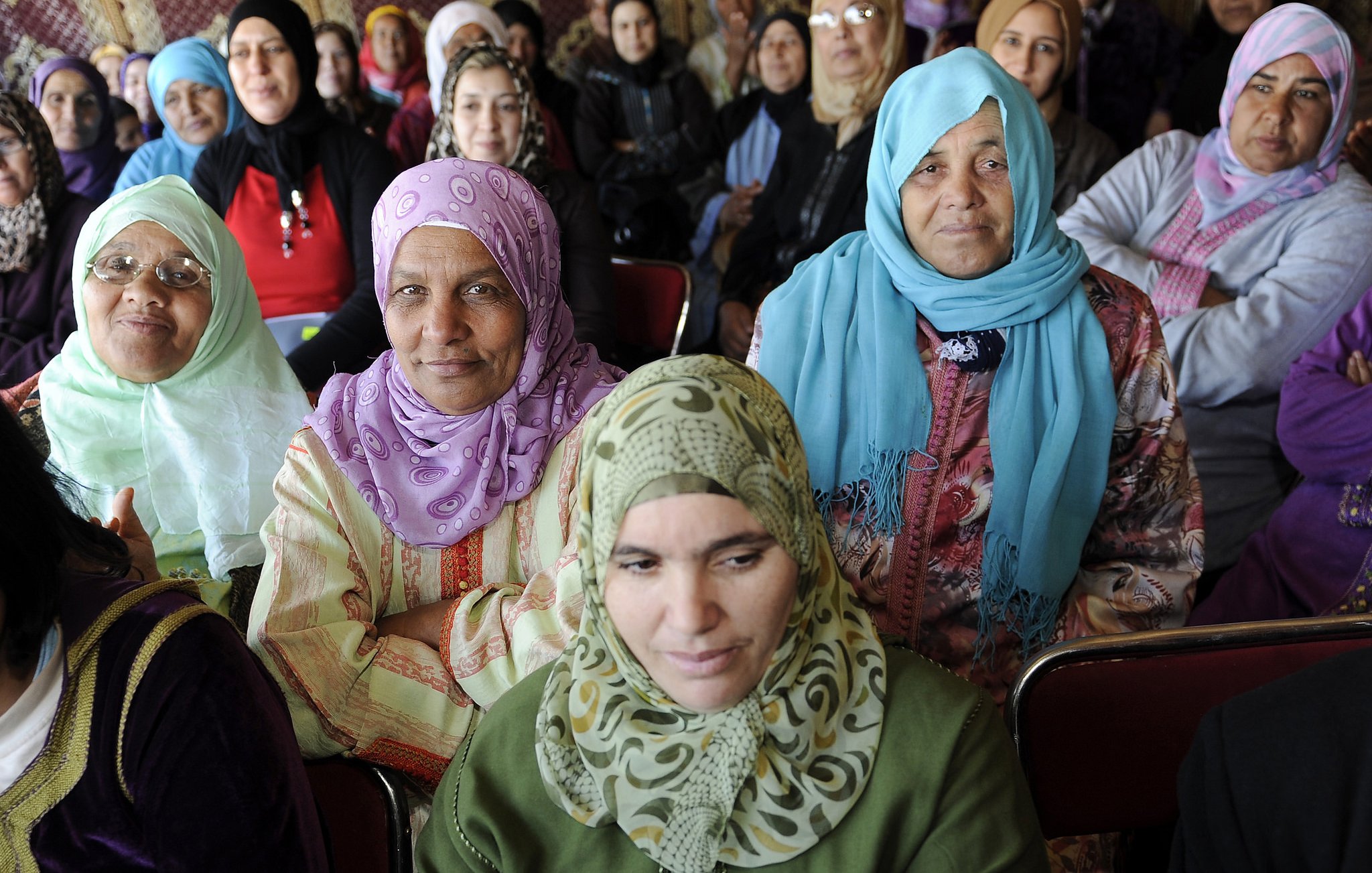 L'image montre un groupe de femmes assises, majoritairement vêtues de vêtements traditionnels et portant des foulards. Certaines portent des vêtements clairs tandis que d'autres ont des tenues plus sombres. Les expressions des femmes varient, allant de souriantes à attentives. L'environnement semble être une réunion ou un rassemblement, avec des murs ornés de motifs. L'atmosphère semble conviviale et solennelle.