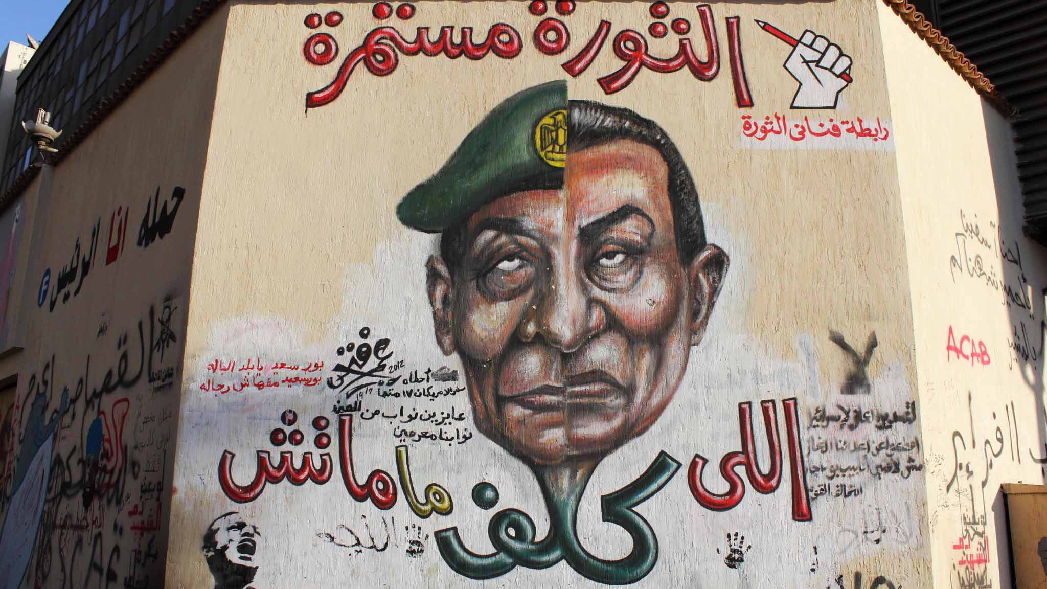 L'image montre une fresque murale sur un mur, représentant un homme avec un uniforme militaire et un képi. Son expression est sérieuse et emblématique. Autour de cette image, on trouve des inscriptions en arabe, dont certaines font allusion à une continuité de la révolution. Le style artistique est vibrant et engagé, évoquant des thèmes de soulèvement et de résistance. Des symboles de révolte ou de mouvement peuvent également être présents, soulignant un message politique fort.