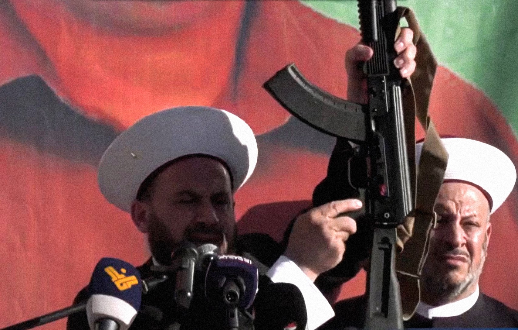 The image depicts two men, wearing white turbans and traditional garments, standing at a podium with microphones. One man is prominently holding an assault rifle above his head, while the other looks on. The background features a large, colorful mural or poster, possibly of a political figure or symbol. The scene suggests a demonstration or rally, conveying themes of power and assertiveness.