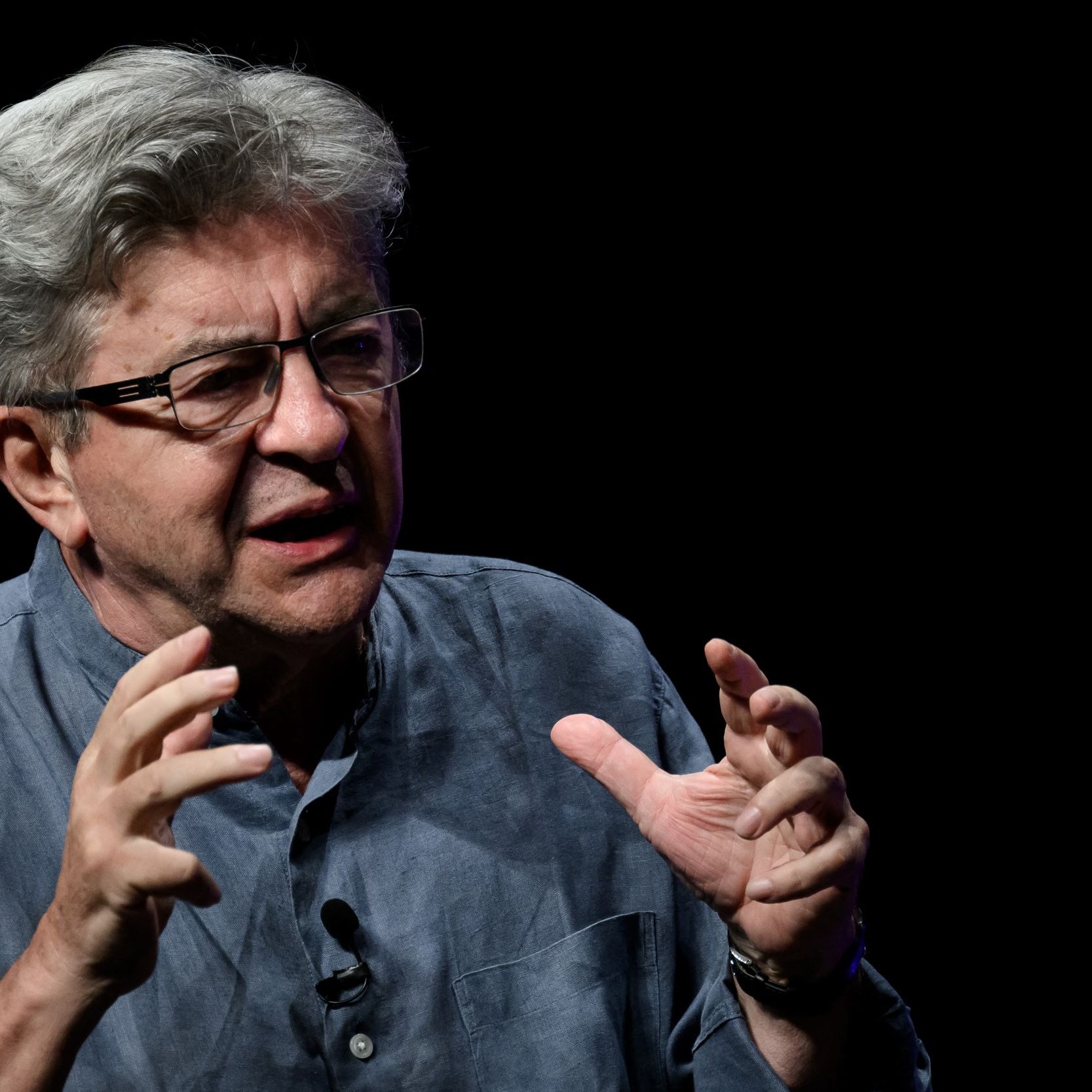 این تصویر یک مرد میانسال را نشان می‌دهد که به نظر می‌رسد در حال صحبت کردن یا بحث کردن است. او عینک به چشم دارد و موهای خاکستری و حالت نامنظمی دارد. لباس او به رنگ آبی تیره یا خاکستری است و به نظر می‌رسد که در یک محیط تاریک یا با پس‌زمینه‌ای سیاه قرار دارد. او دست‌هایش را در حالت ابراز احساسات یا تأکید بر نقاط صحبت خود حرکت می‌دهد.