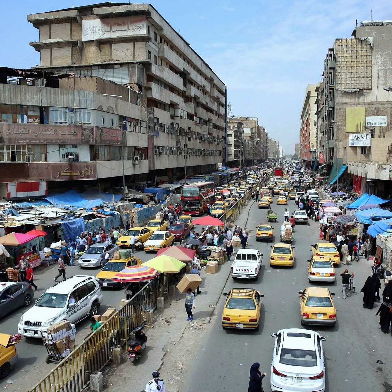 L'image présente une scène urbaine animée, probablement dans une grande ville. On peut voir une rue bordée de bâtiments qui semblent vieux et usés. La circulation est dense, avec de nombreux taxis jaunes qui circulent dans les deux sens. Des échoppes et des marchés de rue sont visibles, avec des auvents colorés, indiquant une ambiance de commerce local. Les gens marchent le long des trottoirs, et l'ensemble de la scène donne une impression de mouvement et de vie citadine. Le ciel est partiellement nuageux, ce qui suggère un climat chaud.