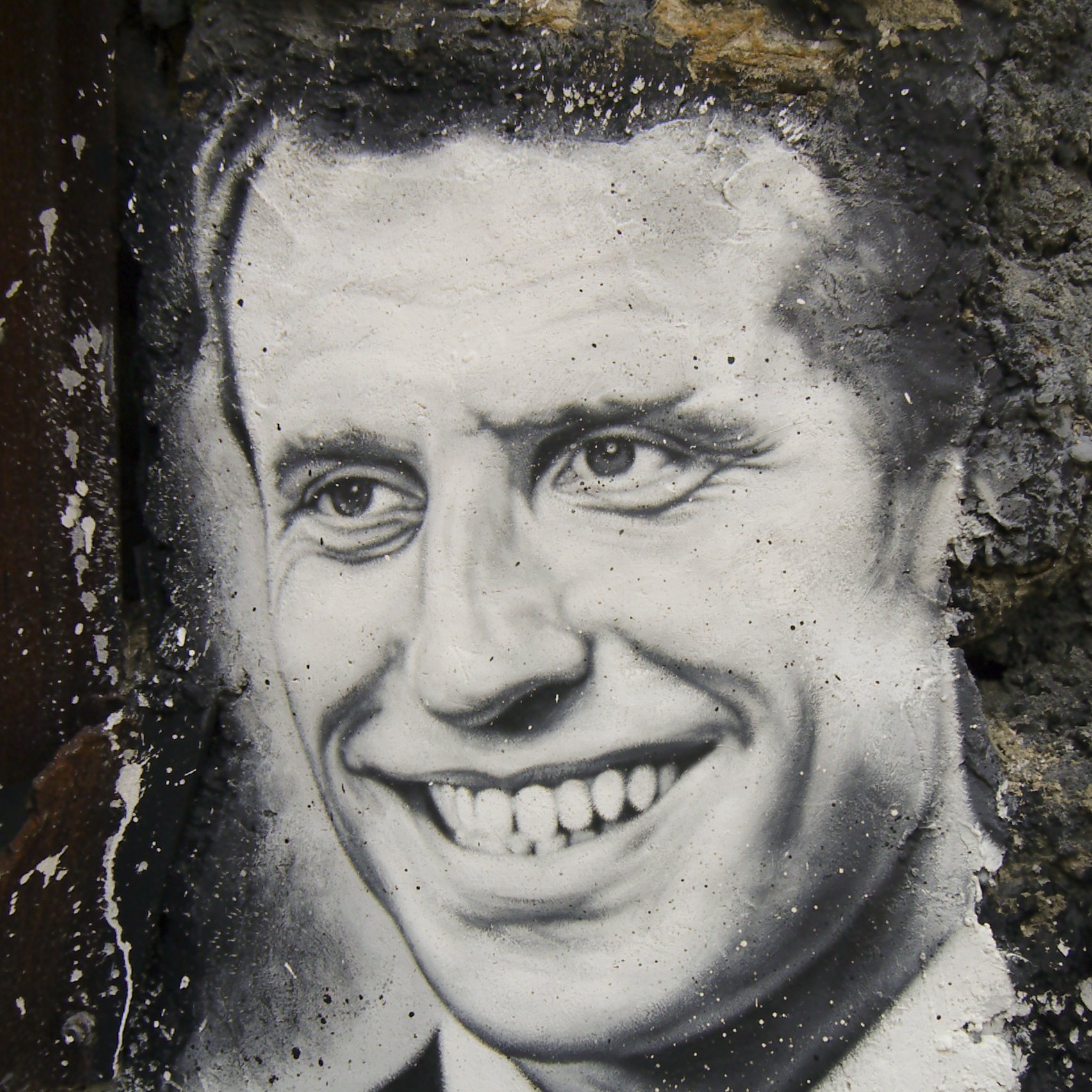 The image features a black-and-white mural of a smiling man. The artwork is set against a textured, stone wall, which adds depth to the scene. The man's expression conveys warmth and friendliness, with a prominent smile and bright eyes. The details are finely rendered, showcasing the artist's skill in capturing facial features and expressions.