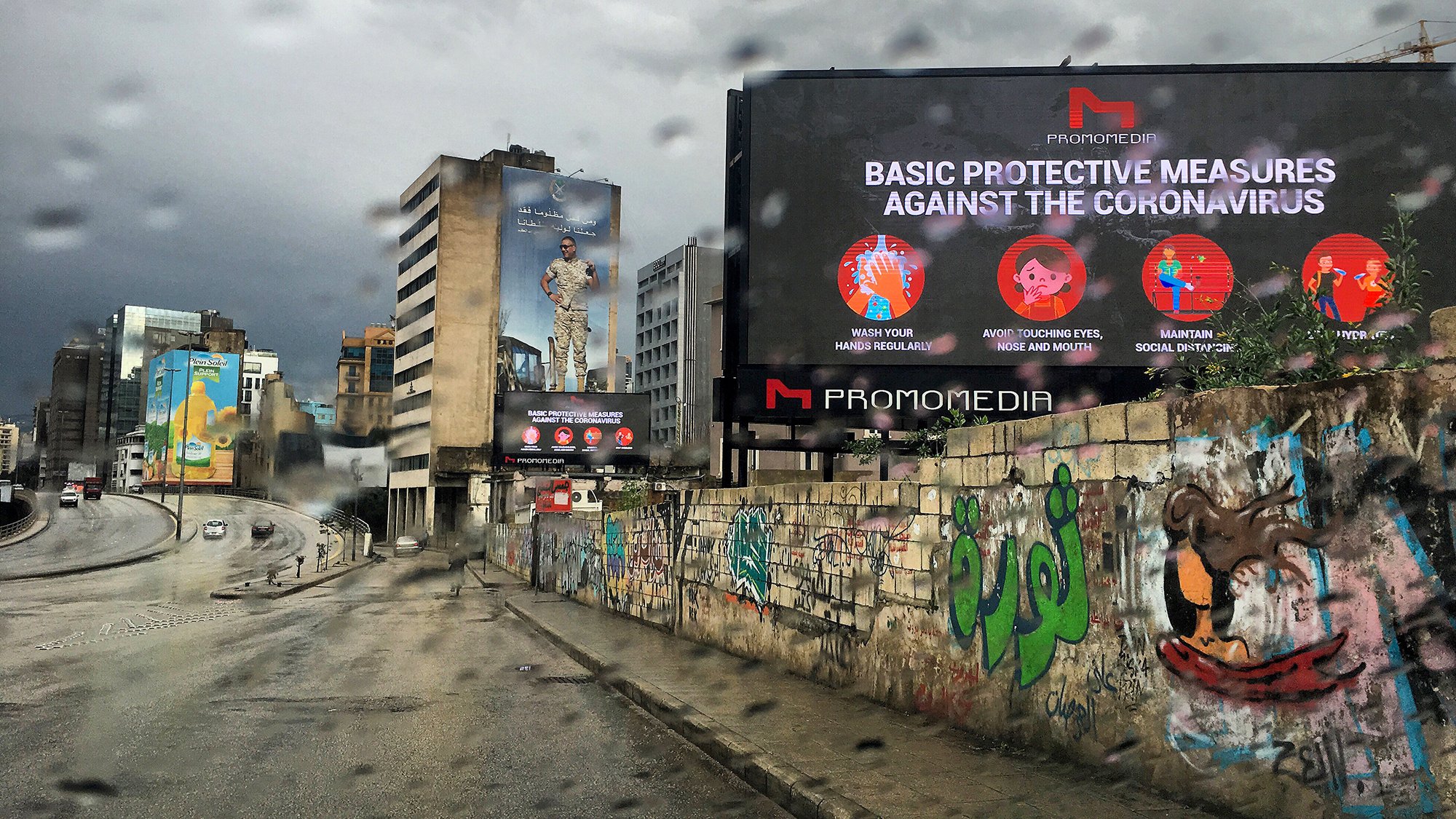 L'image montre une scène urbaine sous la pluie avec des bâtiments modernes à l'arrière-plan. On peut voir un panneau d'affichage numérique qui présente des mesures de protection contre le coronavirus, avec des icônes illustratives. Le mur à gauche est couvert de graffitis colorés, ajoutant une touche artistique à l'environnement. L'atmosphère est plutôt sombre en raison du temps nuageux et pluvieux.