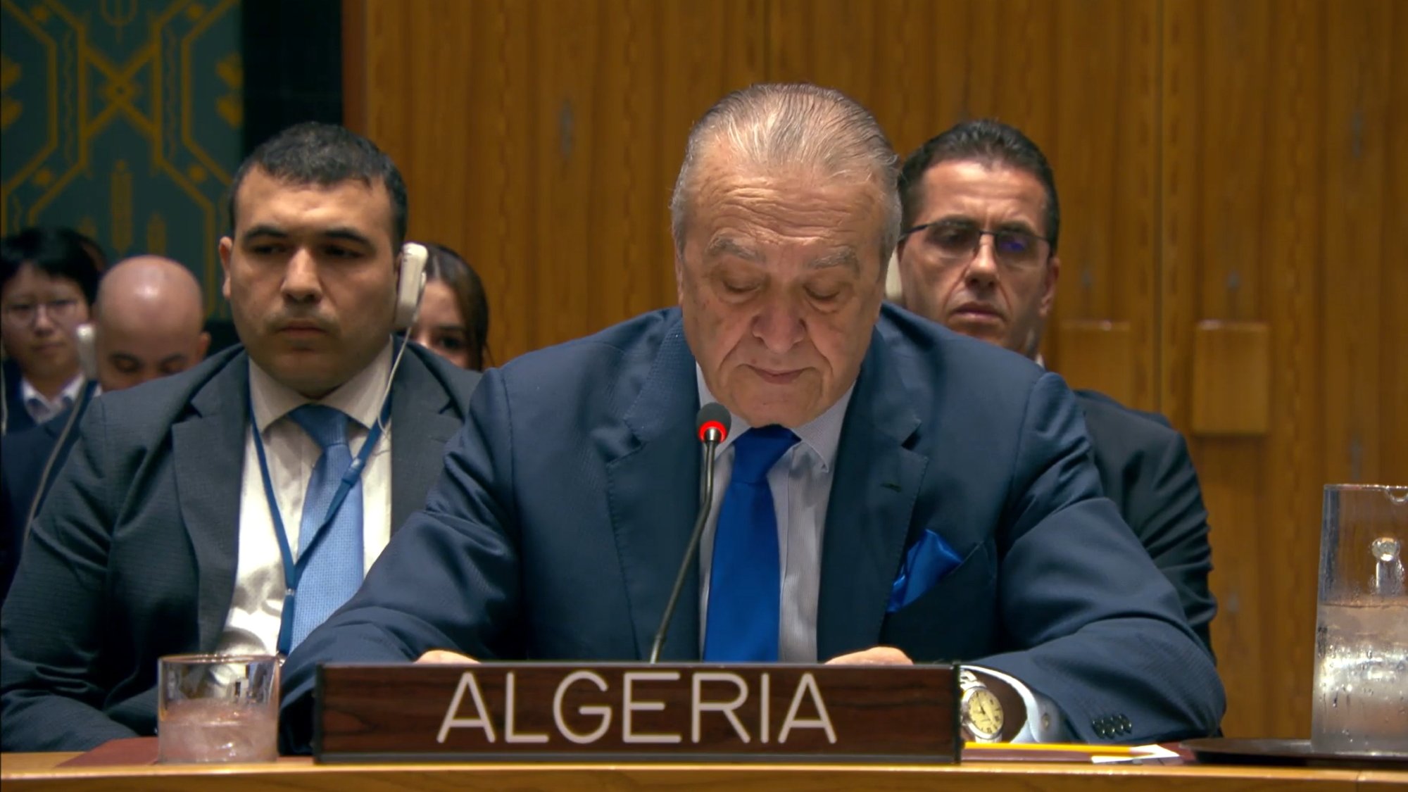 رجل يتحدث خلف منصة، يحمل لافتة الجزائر، مع أشخاص يجلسون خلفه في قاعة.