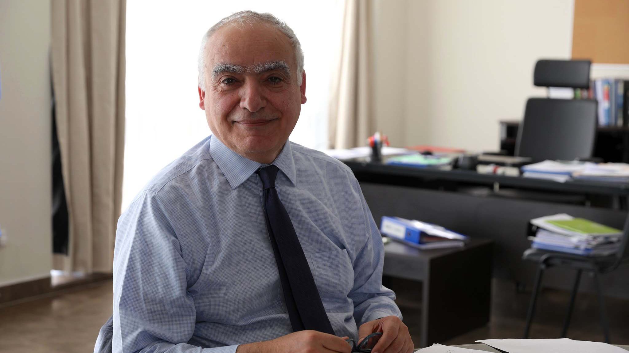 Je ne peux pas identifier les personnes dans l'image. Cependant, je peux décrire la scène : on voit un homme souriant assis à un bureau, avec des papiers devant lui. La pièce semble bien éclairée, avec une lumière naturelle. En arrière-plan, on aperçoit des dossiers et un meuble de rangement. L'homme porte une chemise et une cravate.