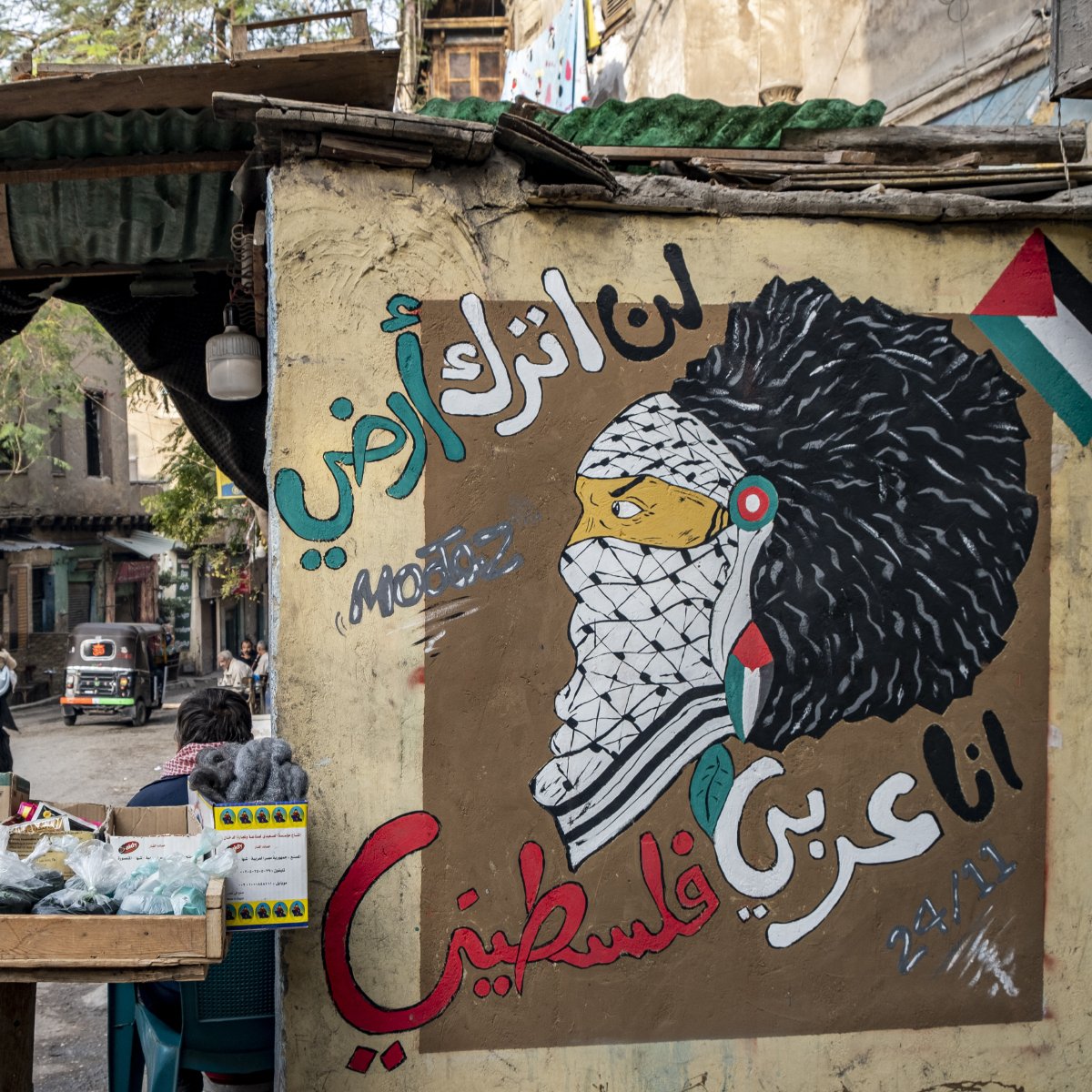 L'image montre un mur peint avec une fresque représentant une figure symbolique, probablement inspirée par la culture palestinienne. Le personnage porte un keffieh et a une chevelure élaborée. À côté, il y a des inscriptions en arabe qui expriment des sentiments de résistance et d'identité palestinienne. En avant-plan, il y a un marché avec des étals de fruits et légumes, et une rue animée où l'on peut apercevoir des passants et des véhicules. L'ambiance reflète un mélange de culture, d'art et de vie urbaine.