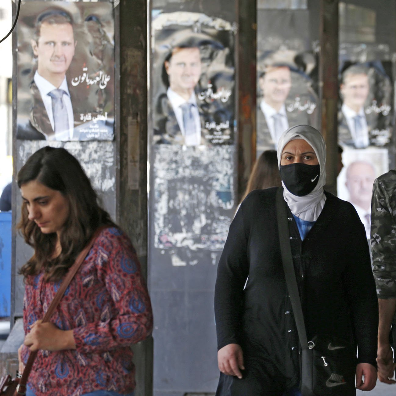 L'image montre une rue animée avec des personnes marchant sous un abri. En arrière-plan, on aperçoit des affiches de propagande, probablement de l'homme politique en question. Plusieurs personnes, dont une femme portant un hijab et un masque, se déplacent le long du trottoir. L'atmosphère semble dynamique et quelque peu urbaine, avec des jeunes et des adultes interagissant dans cet espace public.