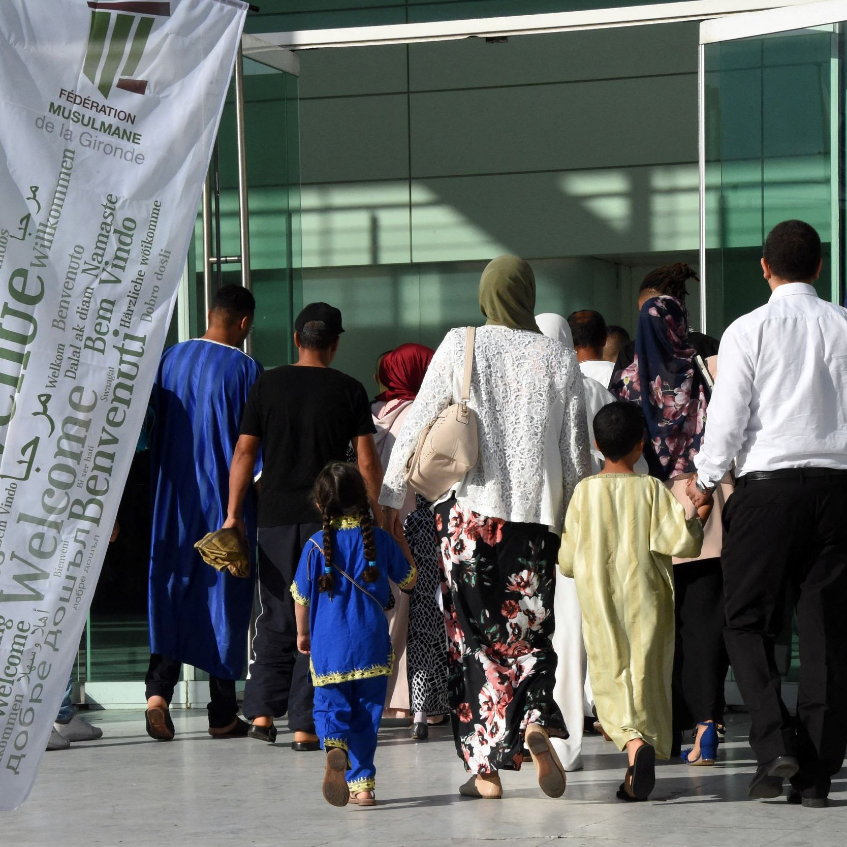L'image montre un groupe de personnes se dirigeant vers un bâtiment. On peut voir des individus de différentes générations, y compris des enfants. Certains portent des vêtements traditionnels, et plusieurs femmes ont des foulards. Il y a également une grande bannière blanche flottant au vent, portant le mot "Bienvenue" en plusieurs langues. L'ambiance semble chaleureuse et accueillante.