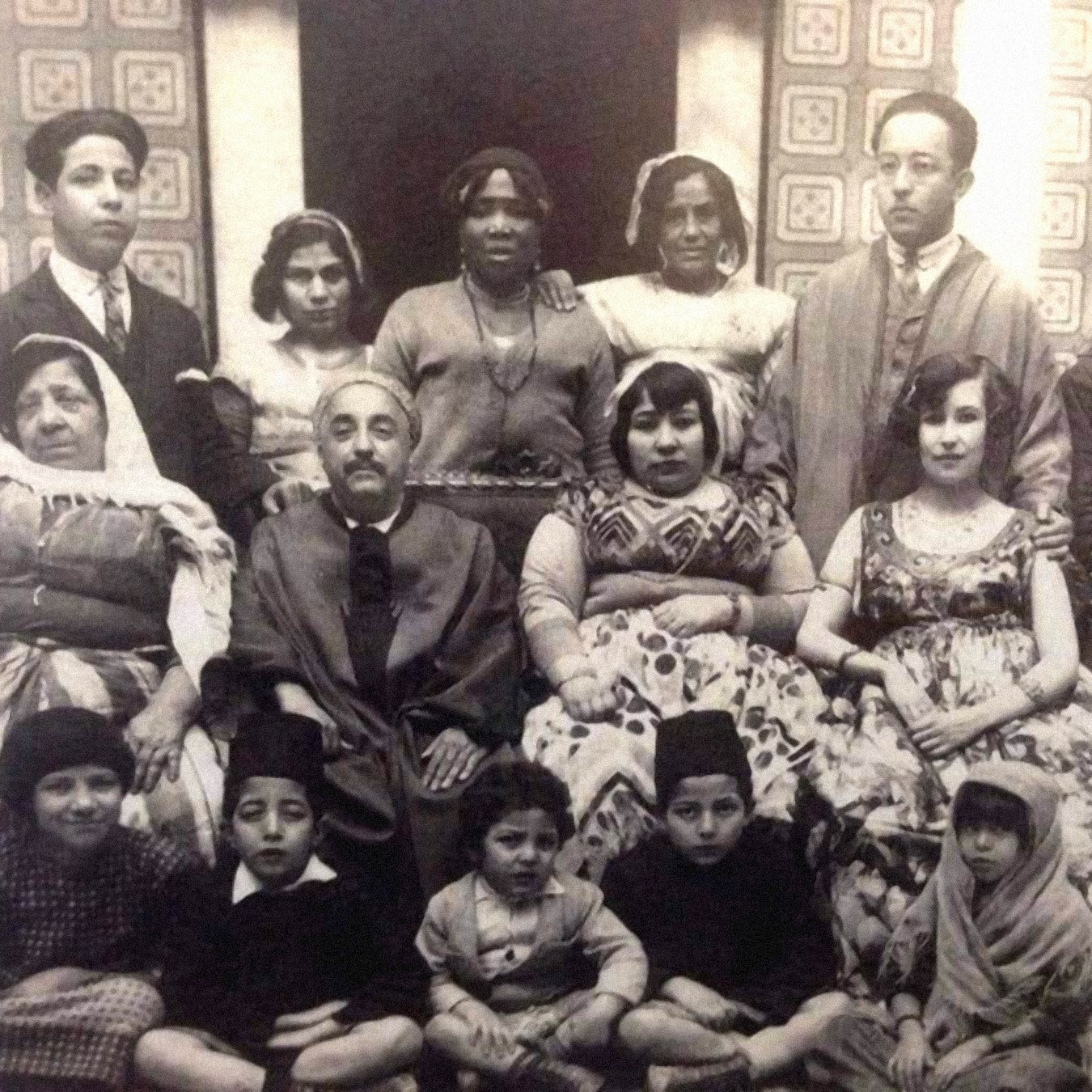 L'image présente un groupe de personnes, probablement une famille ou une communauté, posant ensemble dans un décor intérieur. On peut voir des hommes et des femmes de différentes générations, ainsi que des enfants, vêtus de tenues traditionnelles. L'atmosphère semble conviviale et chaleureuse. L'arrière-plan est orné de motifs, ajoutant une touche historique à la scène.